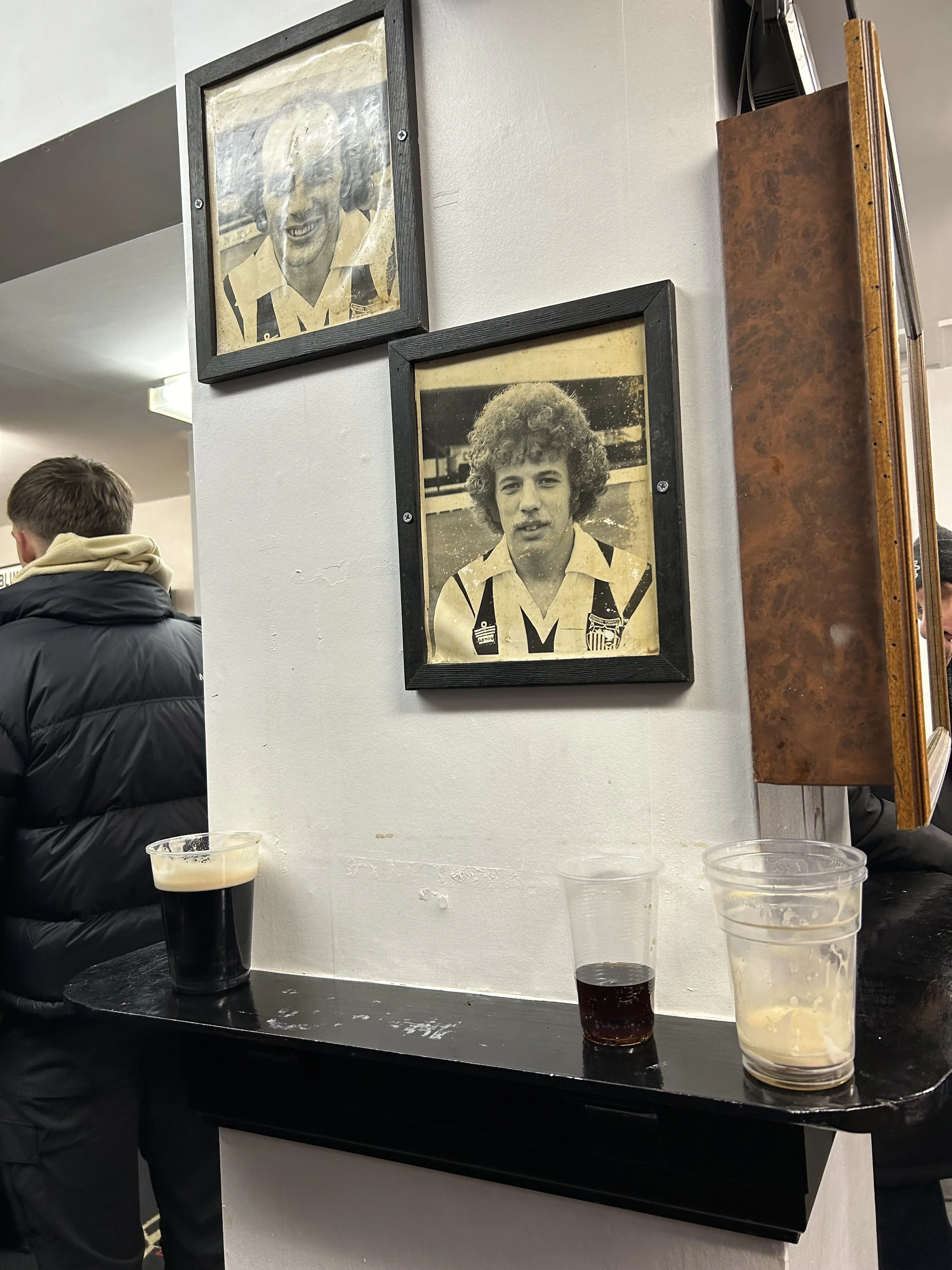 Black and white framed photos of two soccer players, one with curly hair and another with a smiling face, on a white wall behind a black shelf with drinks on it, in a busy indoor setting.
