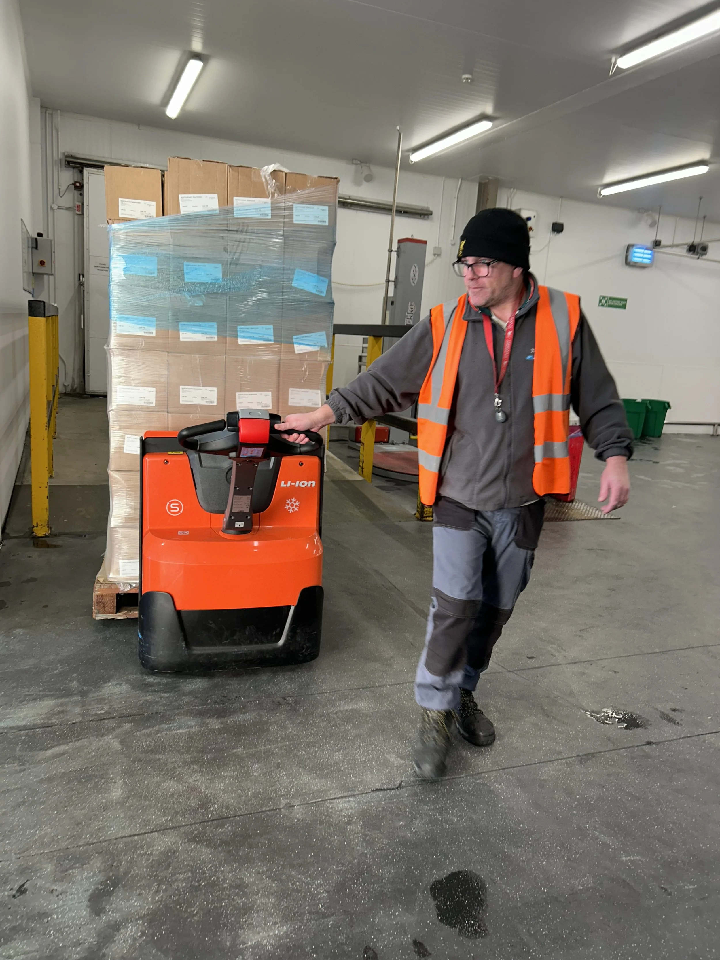 A man wearing a black beanie, glasses, and an orange safety vest is pushing an orange lithium-ion battery-powered industrial pallet jack in a warehouse, with a pallet of boxes in the background.