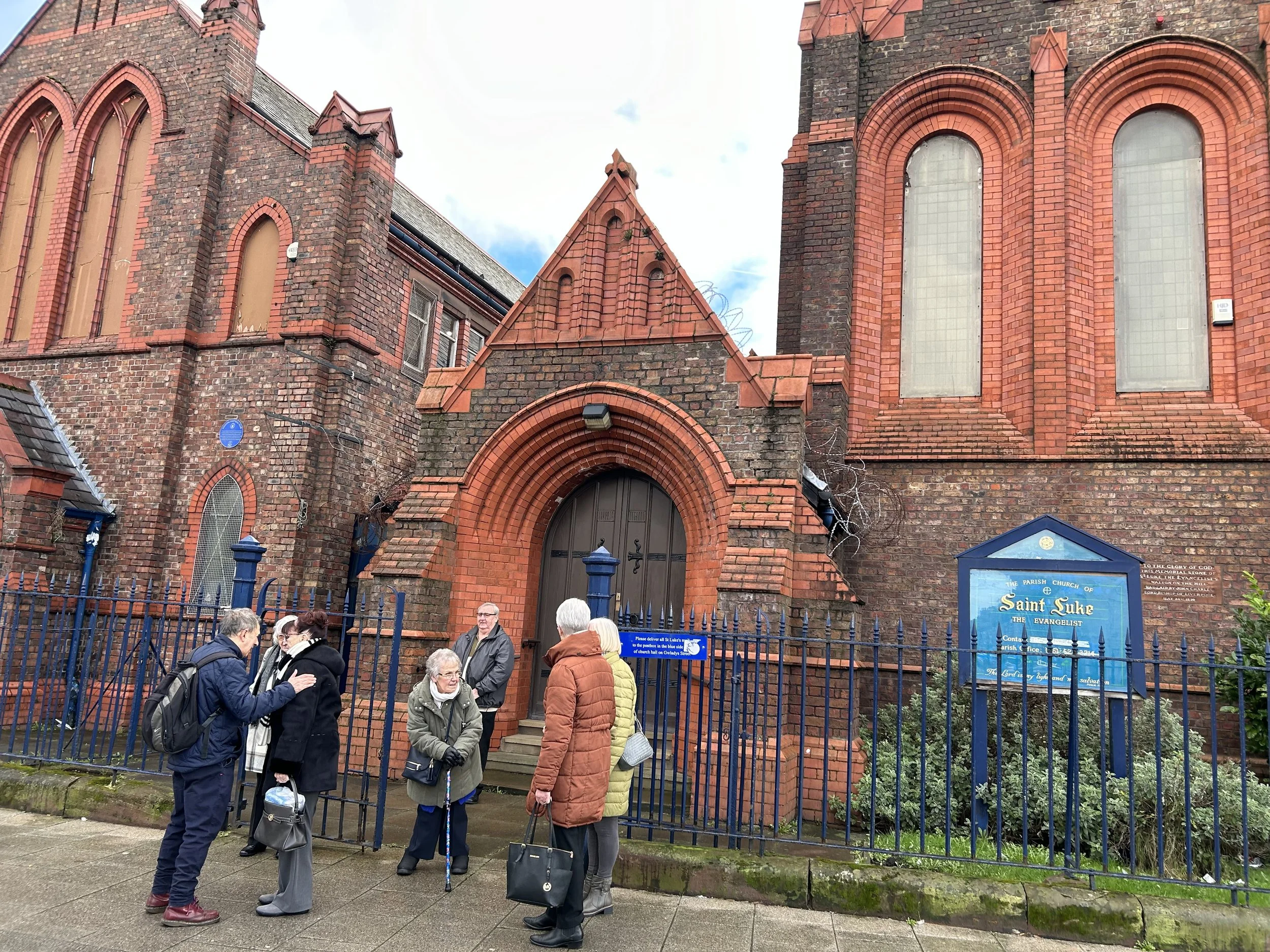 A group of people gathered outside a red brick church, Saint Luke, with high arched windows and a blue sign, on a cloudy day.
