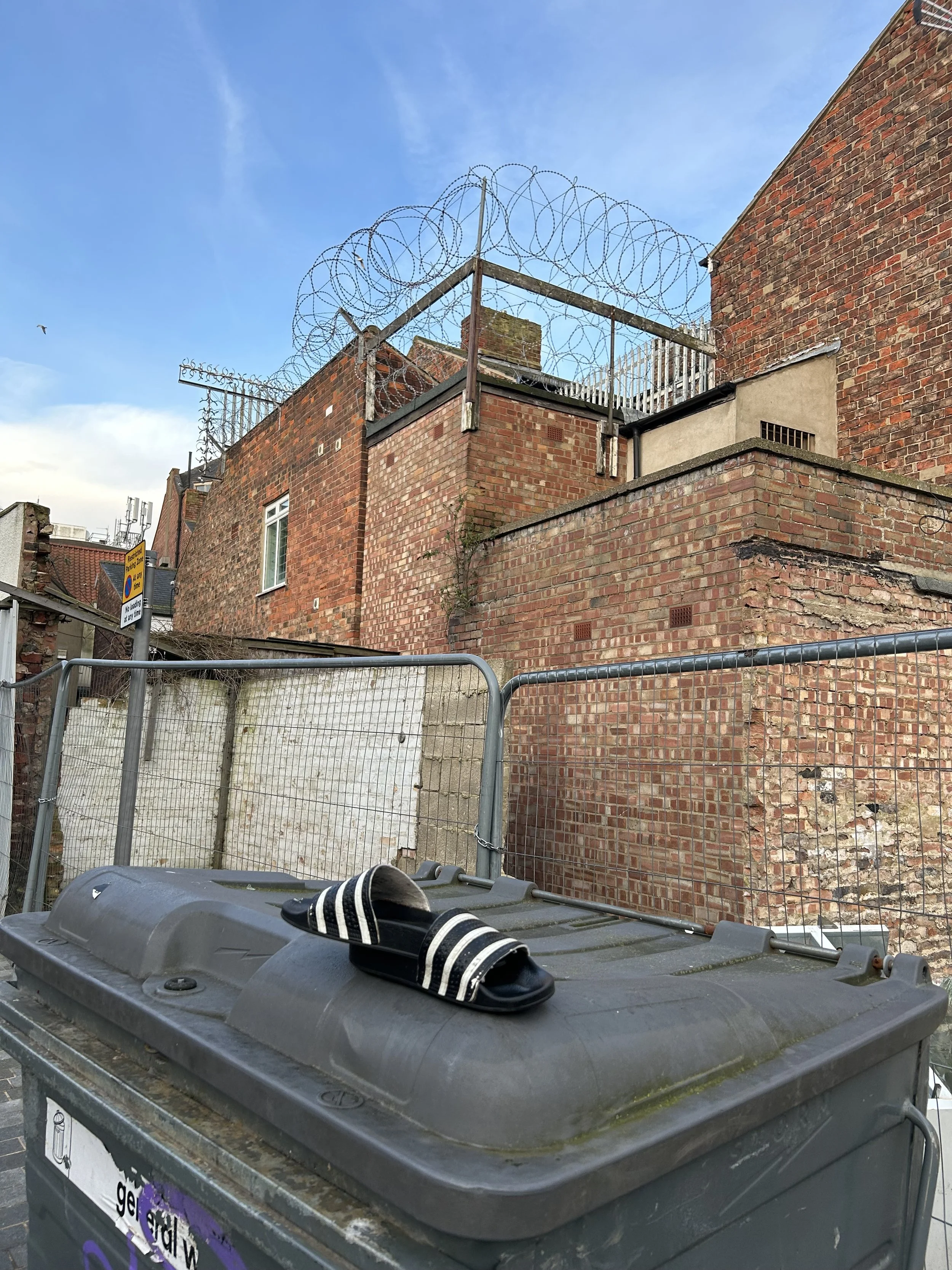 A pair of black and white striped slide sandals resting on a gray wheelie bin in an urban alleyway with brick buildings, a metal fence, and barbed wire on top of a brick wall against a blue sky.