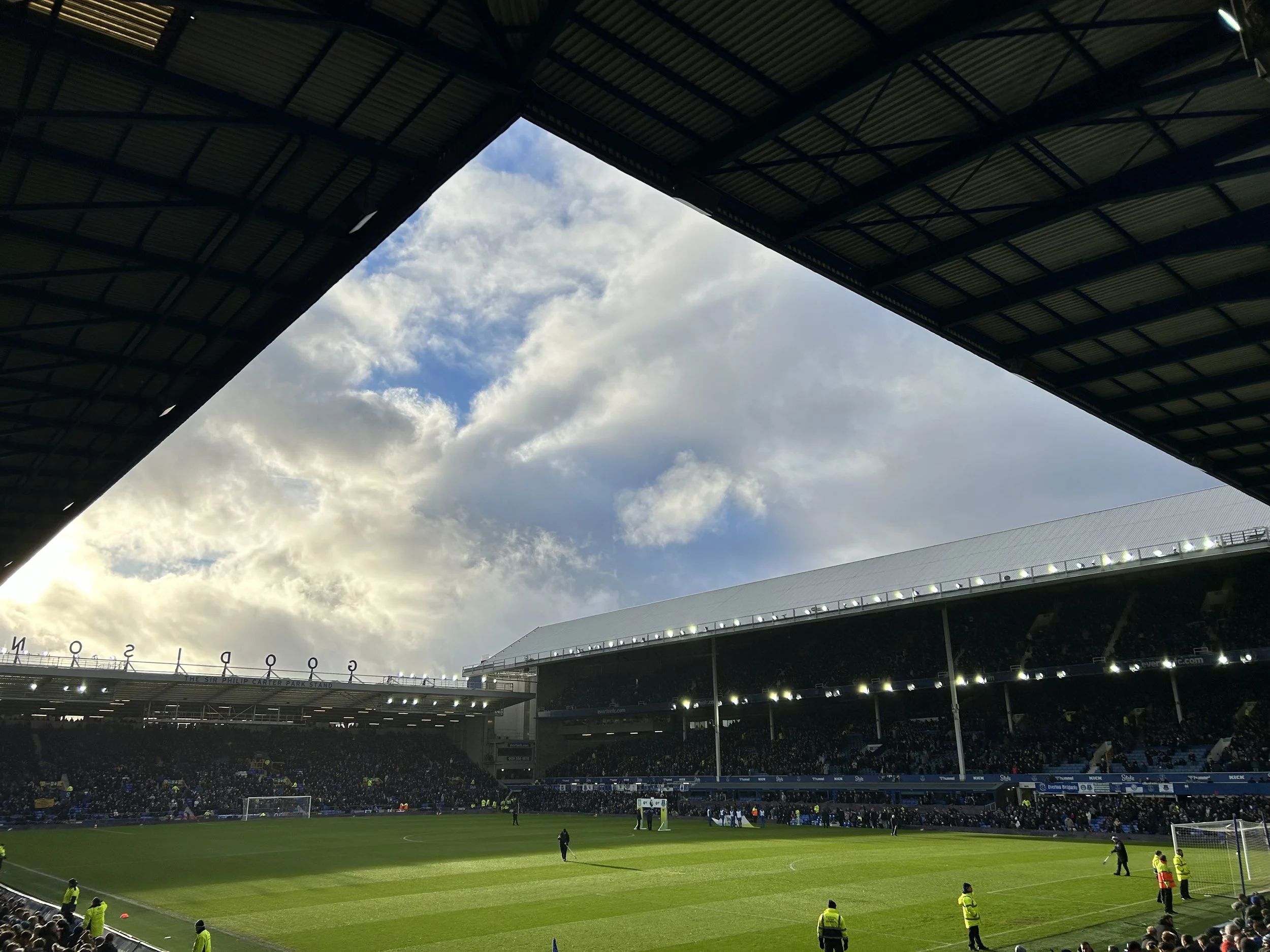 View of a stadium with a partially covered roof, a large crowd of spectators, and a green football field with players and security personnel.