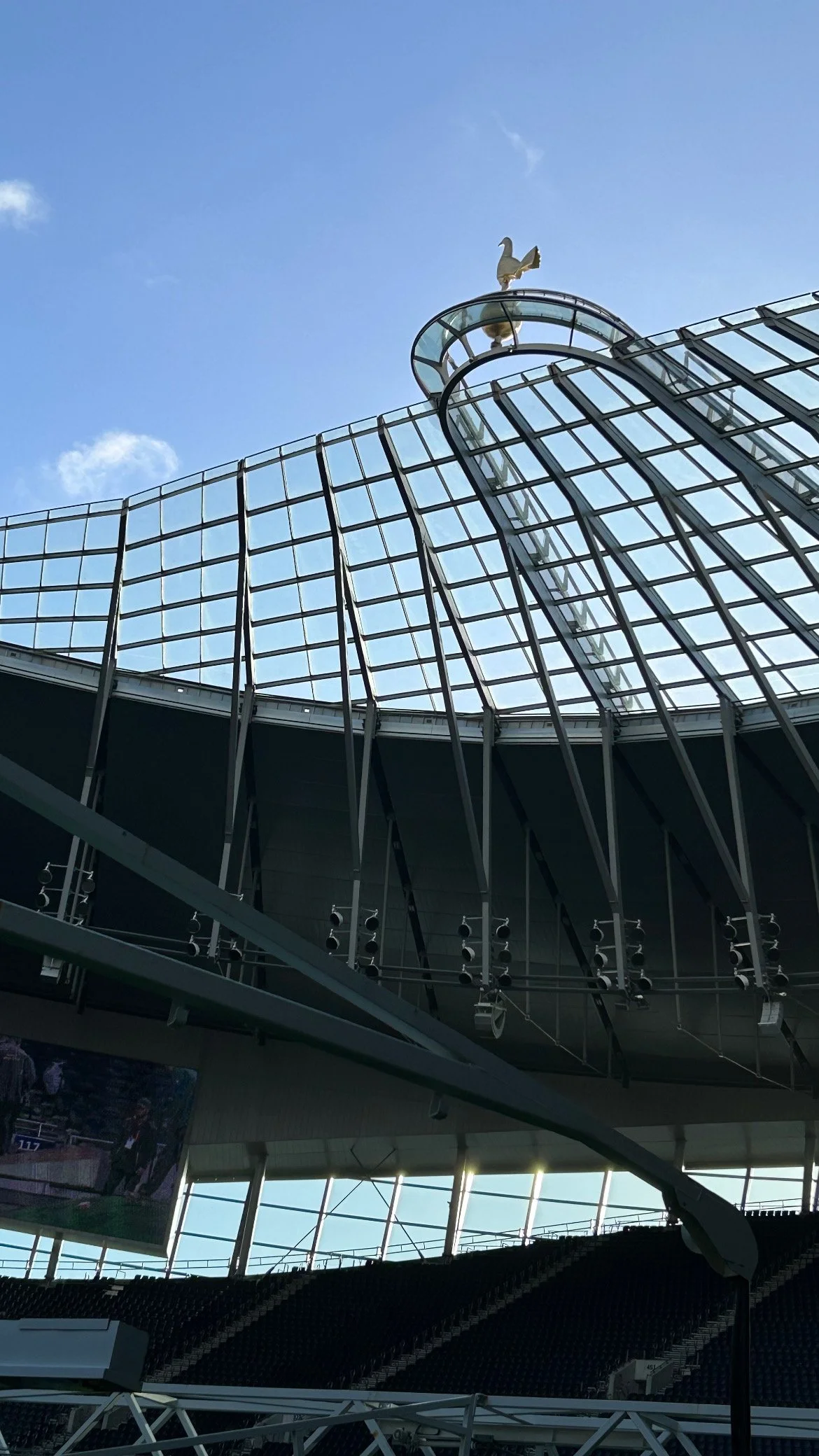 A modern stadium with a glass roof structure and a decorative weather vane in the shape of a rooster on top, viewed from inside.