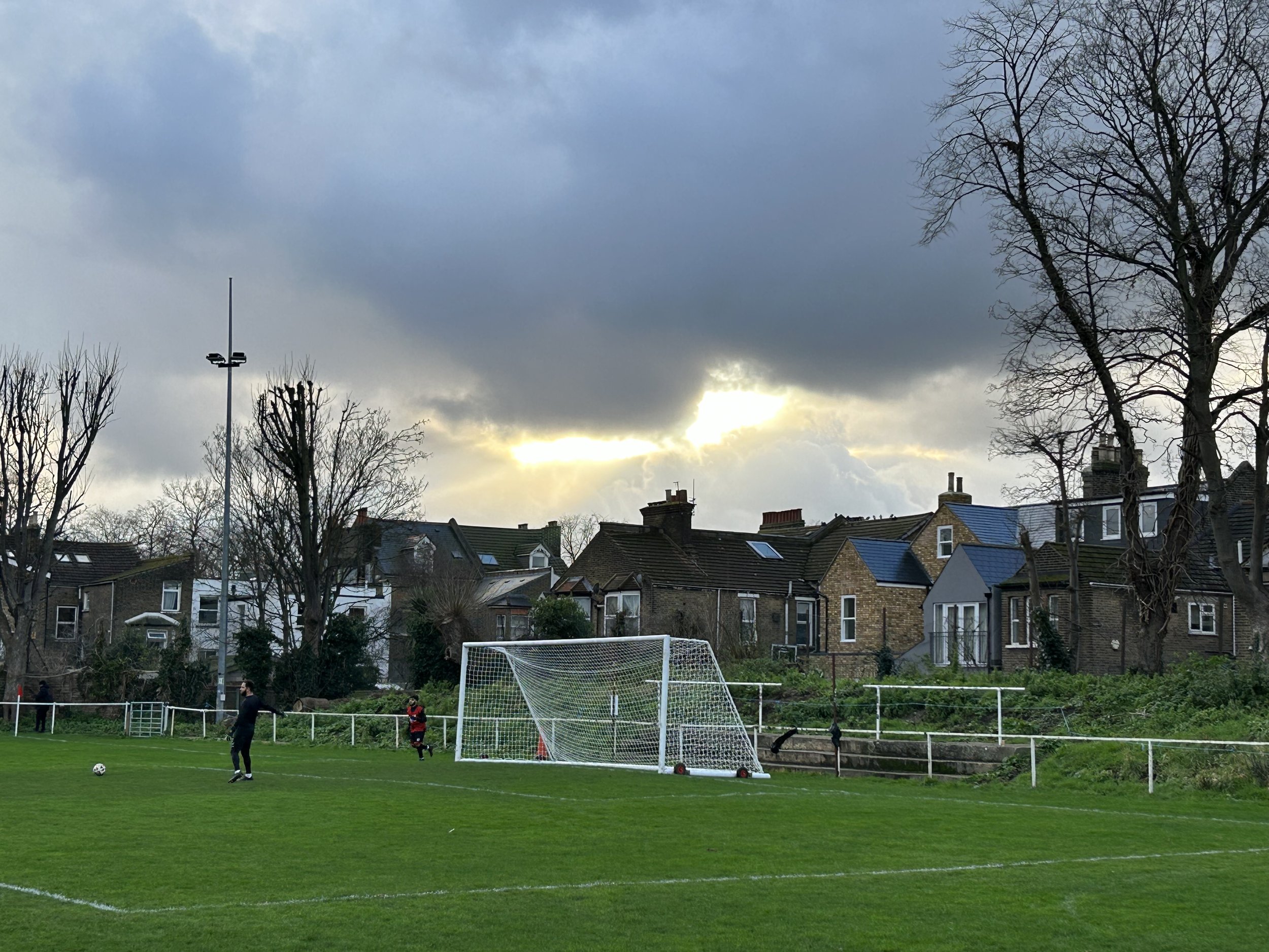 A soccer field with a goalpost and a person in a black jacket and a child in a red and black outfit near it. In the background, there are residential houses and leafless trees, with a cloudy sky and the sun peeking through.