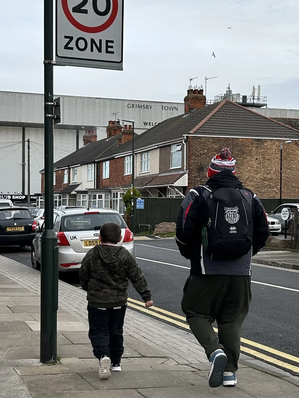 A boy and an adult walking on a sidewalk in front of parked cars, with a street and houses in the background, and a sign indicating a 20 mph speed limit zone.