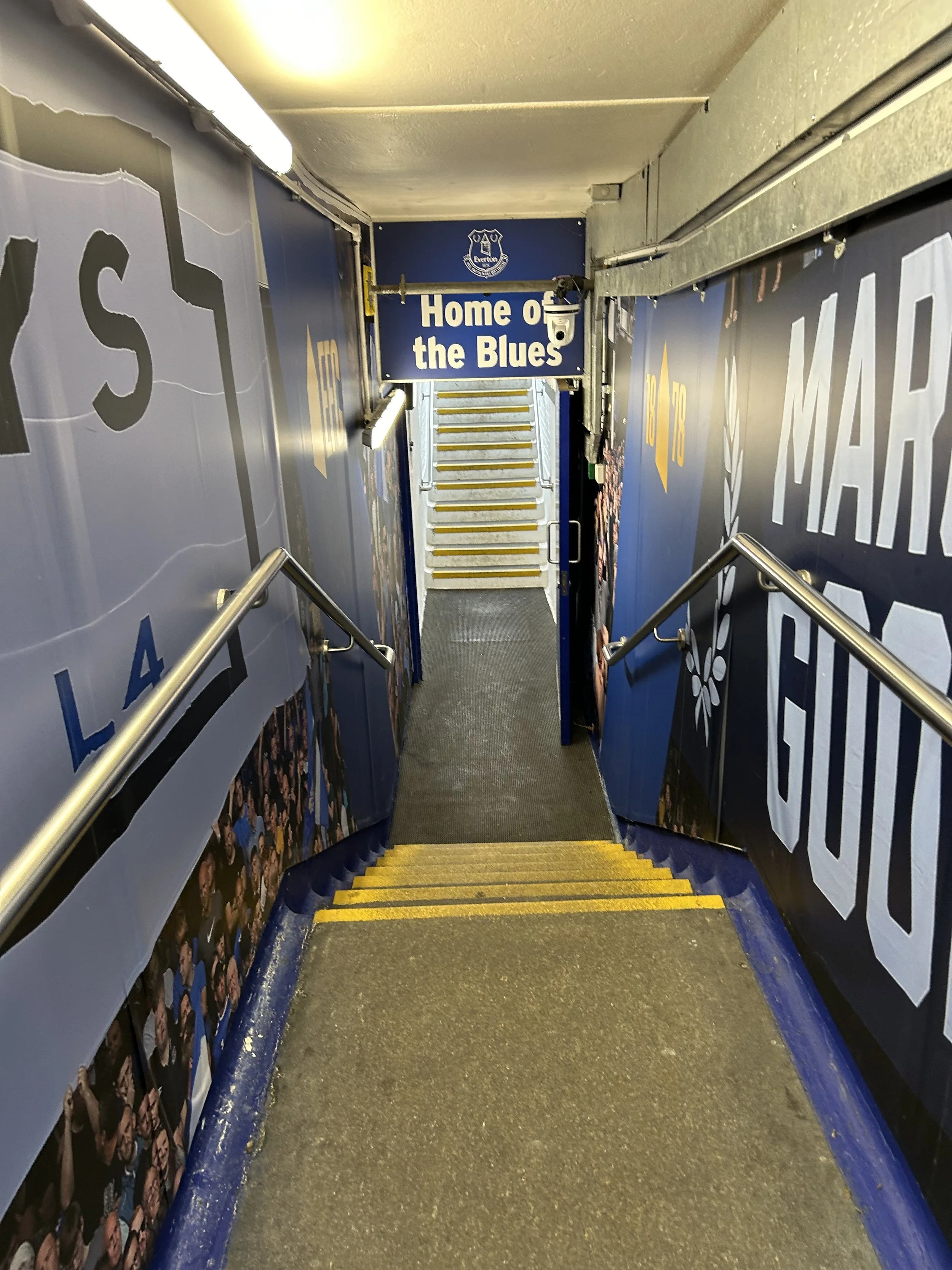 Indoor stadium stairs leading to an area with a sign that reads "Home of the Blues"; blue walls with team logos and advertisements.
