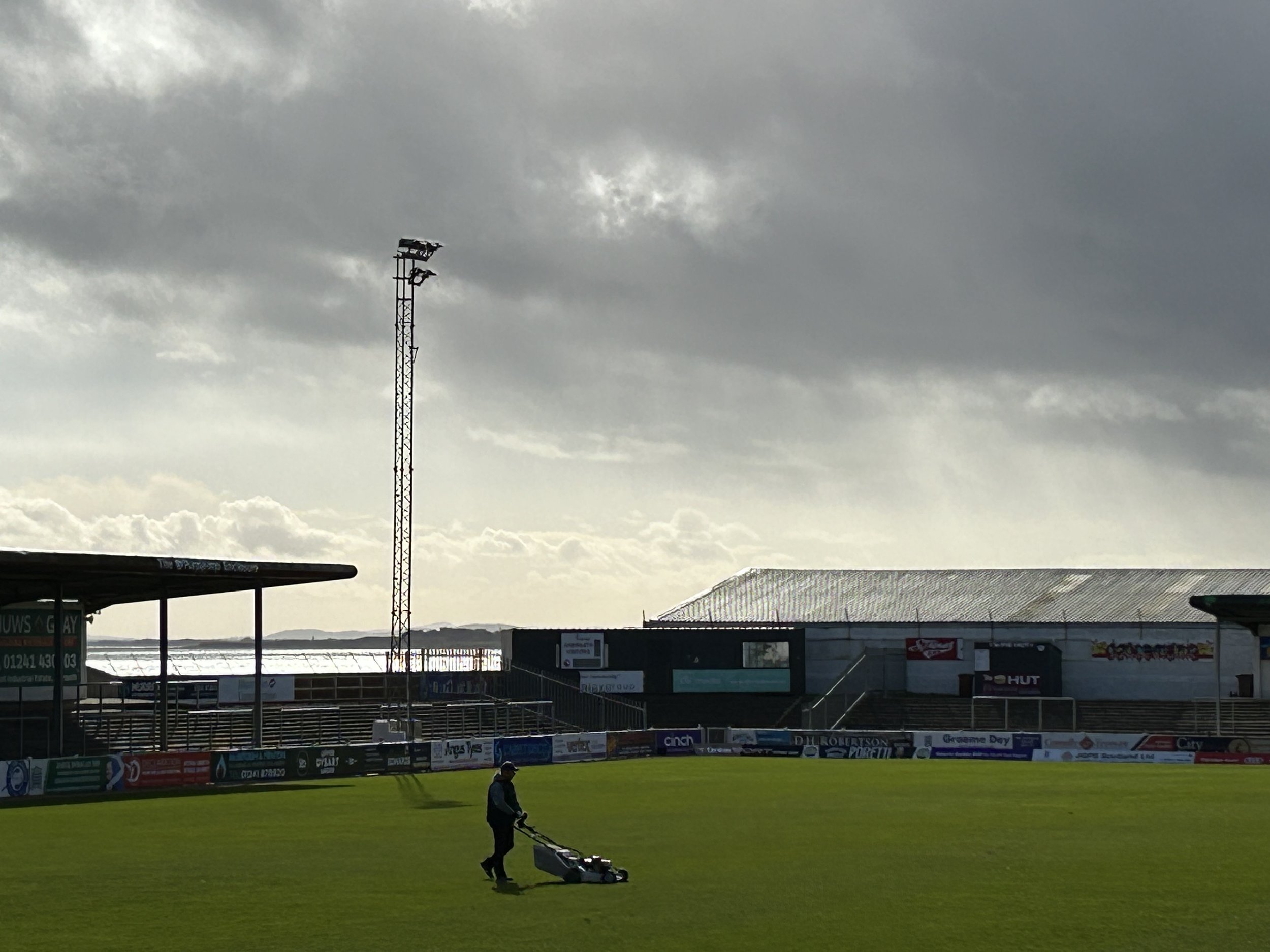 A person mowing the grass on a sports field under cloudy skies, with stadium seating, banners, and a tall floodlight in the background.