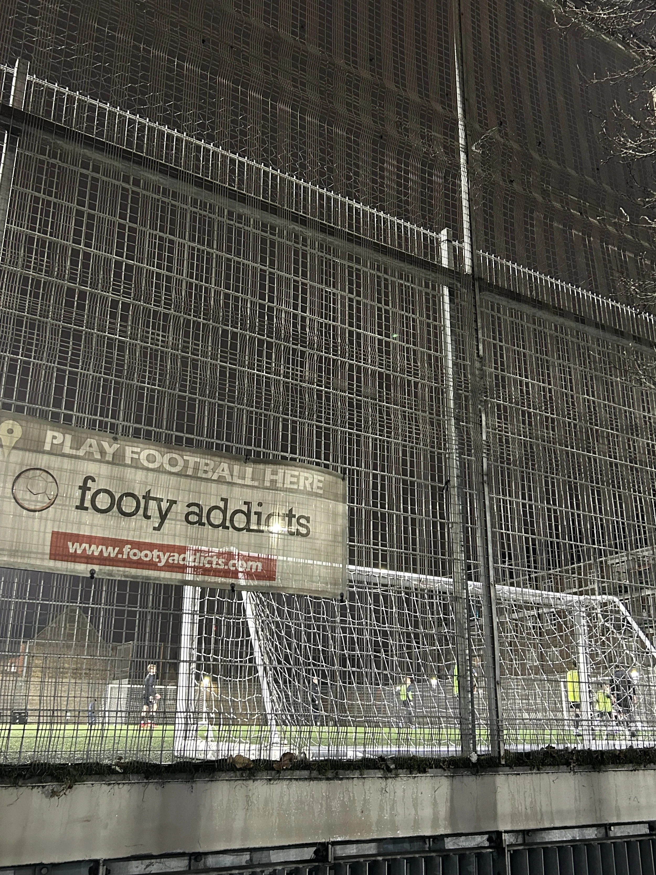 View of a football field seen through a tall metal fence with a sign that reads 'PLAY FOOTBALL HERE' and 'footy addicts' with a website www.footyaddicts.com, with players on the field in the background.