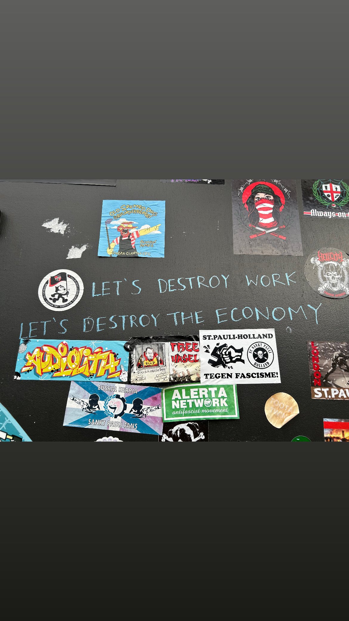 A black surface with various stickers and posters, including a handwritten message in blue ink that says, 'LET'S DESTROY WORK, LET'S DESTROY THE ECONOMY'.