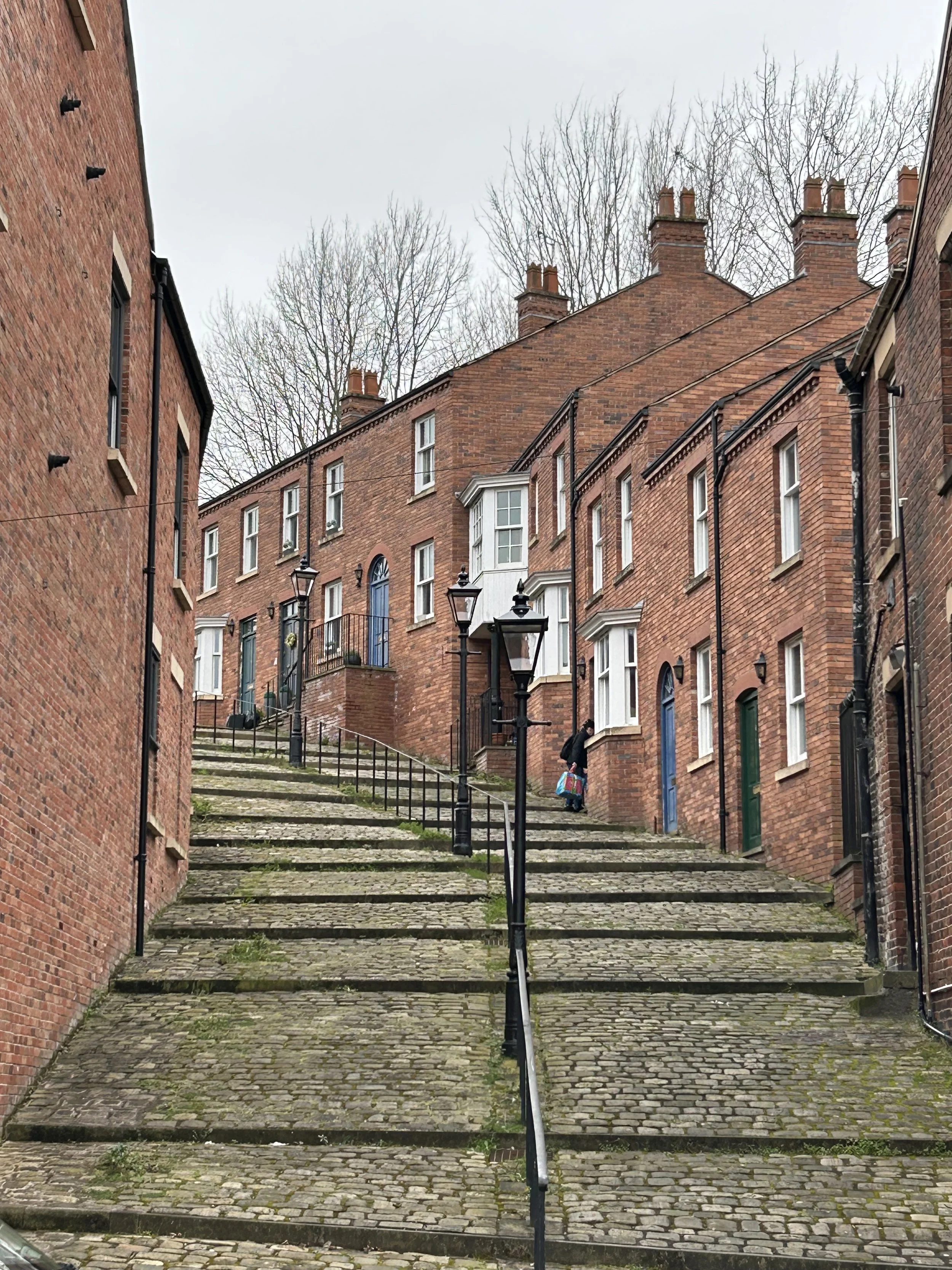 A steep cobblestone street with lansdcaped steps, black railings, and vintage street lamps, leading up to red brick houses with bay windows in an urban neighborhood.
