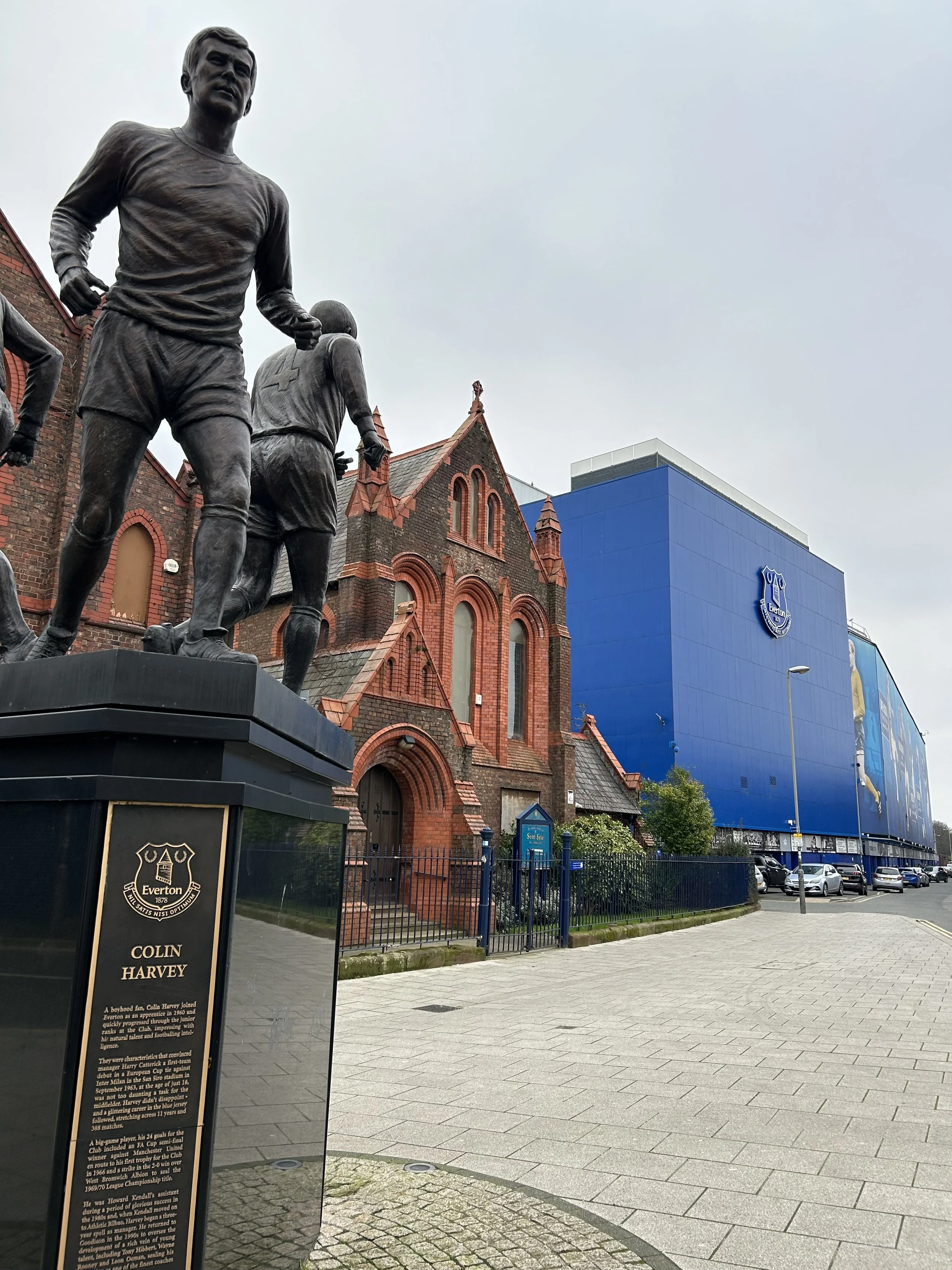 Bronze statue of football player Colin Harvey in front of a red brick church, with modern blue building and parked cars in the background.