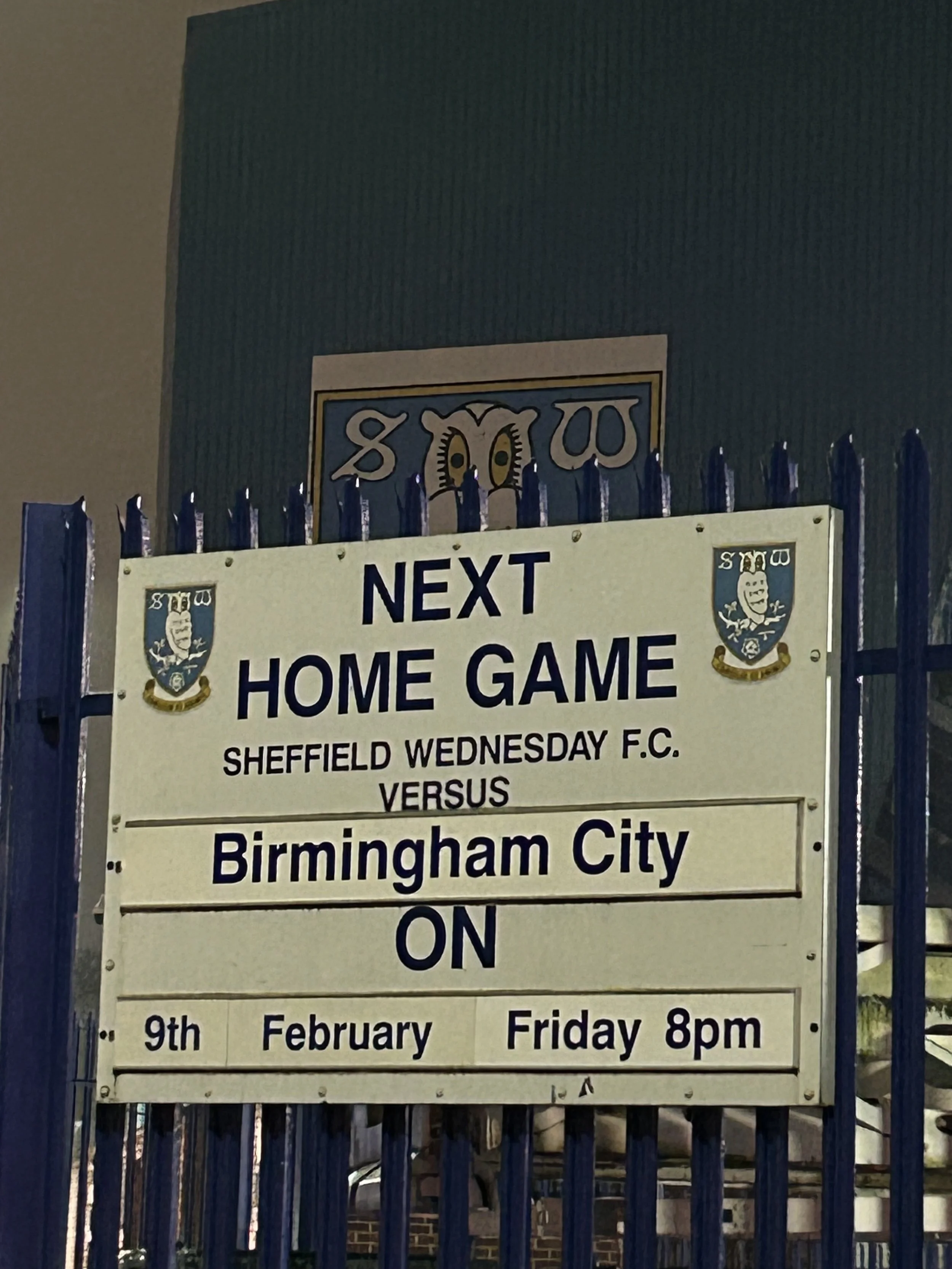 Sign announcing the next home game for Sheffield Wednesday F.C. against Birmingham City, scheduled for February 9th, Friday at 8 pm, posted behind a blue fence with the Sheffield Wednesday logo.