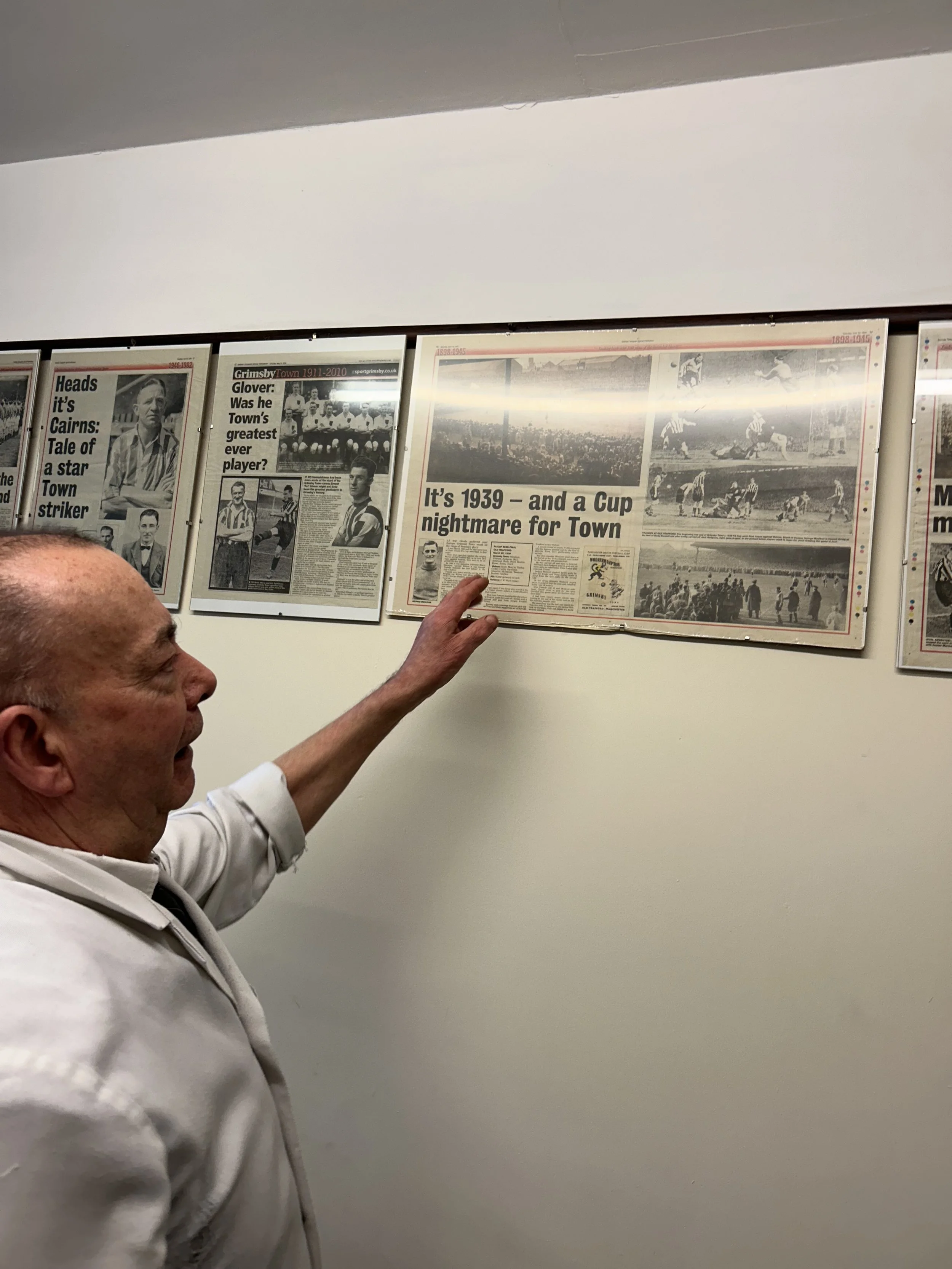 A man in a white jacket points at a newspaper article on a wall display, which features black-and-white photos and headlines about the year 1939 and local football history.