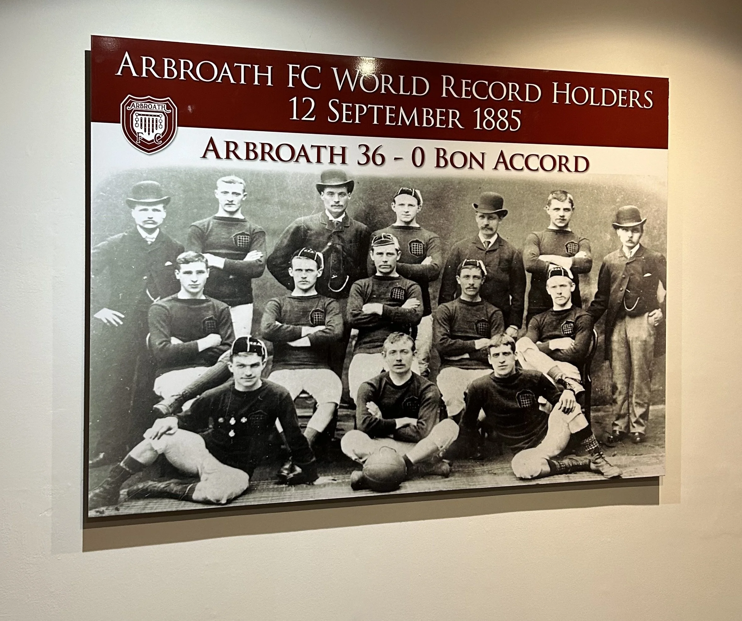 Historical photograph of a football team, Arbroath FC, from 1885, with players in vintage uniforms, some sitting and some standing, and a sign with text about their record.