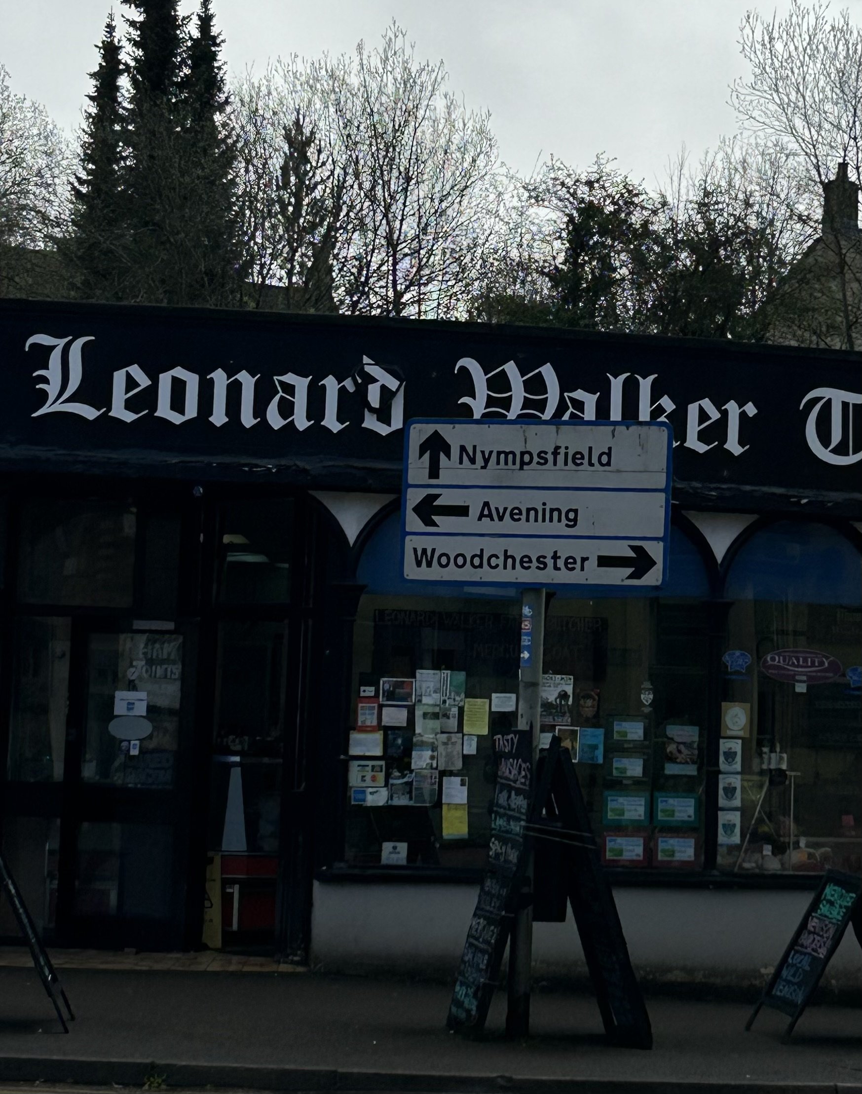 Street view of Leonard Walker store with directional road signs pointing to Nympsfield, Avening, and Woodchester, in front of a building with notices in the window and trees in the background.
