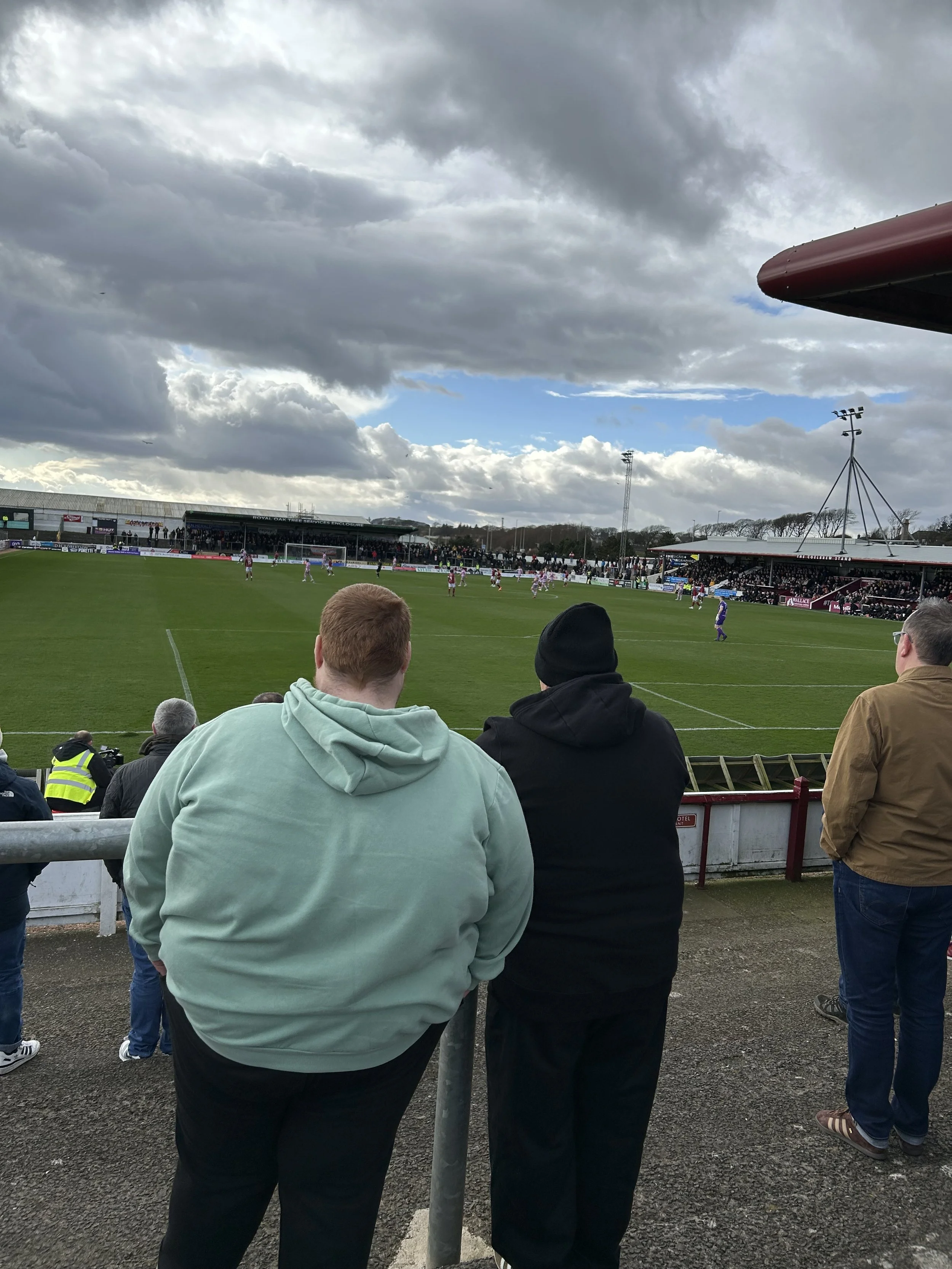 People watching a football game at a stadium with dark clouds overhead and a partly cloudy sky.