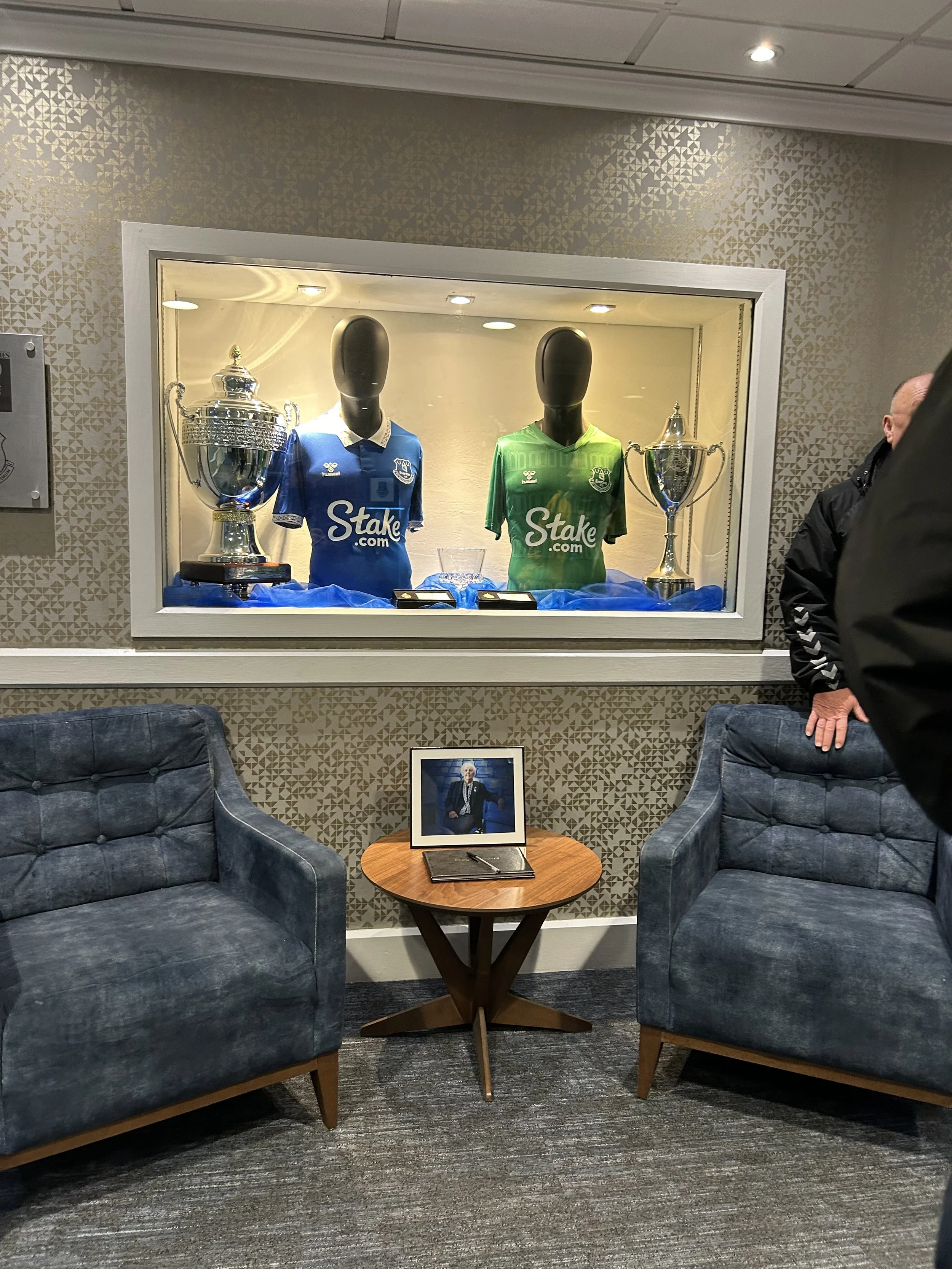 Display of two football jerseys, one blue and one green, each with a trophy beside it, in a showcase with blue fabric underneath. A framed photo of a man in a suit on a small round table in front of two blue armchairs. A person in a black jacket part