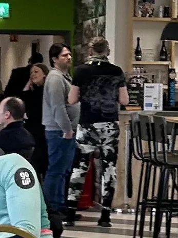 People standing and chatting inside a coffee shop or cafe, with shelves and bar items in the background.