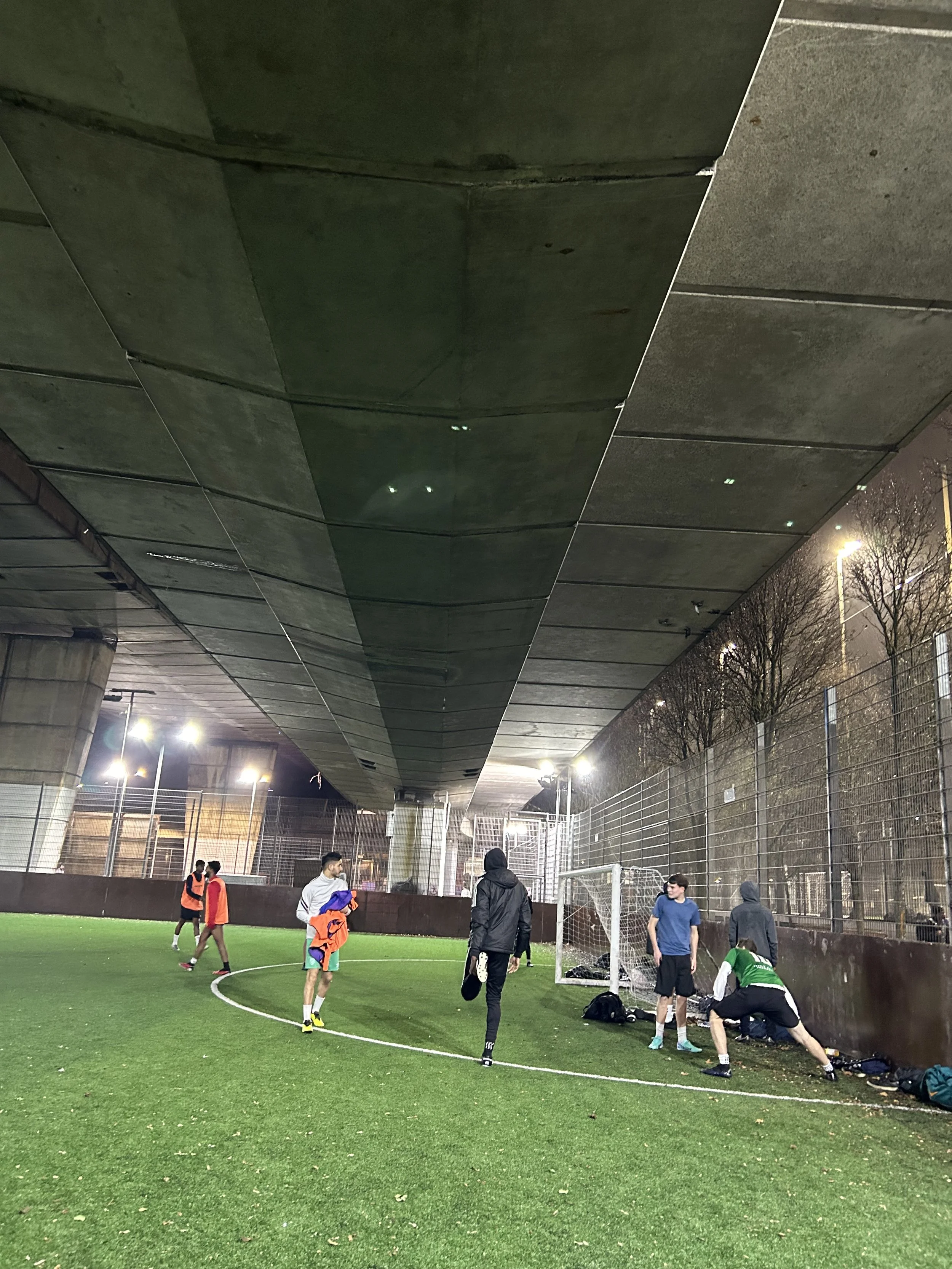 Group of young men playing soccer on an artificial turf field under a bridge at night.