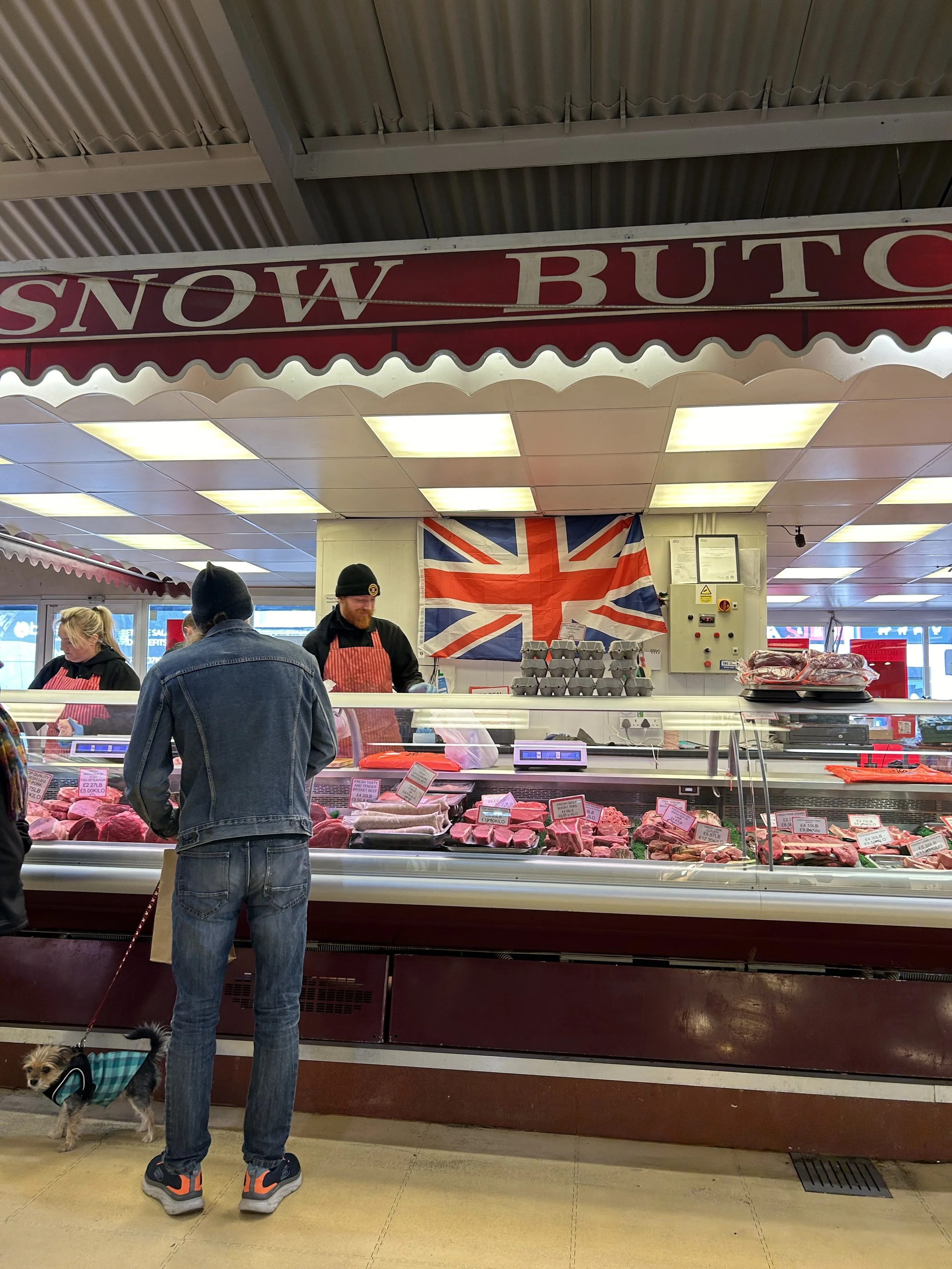 A man with a dog shopping at a meat counter in a British-themed grocery store, with a Union Jack flag hanging on the wall behind the counter, above a sign reading 'Snow Butc'.
