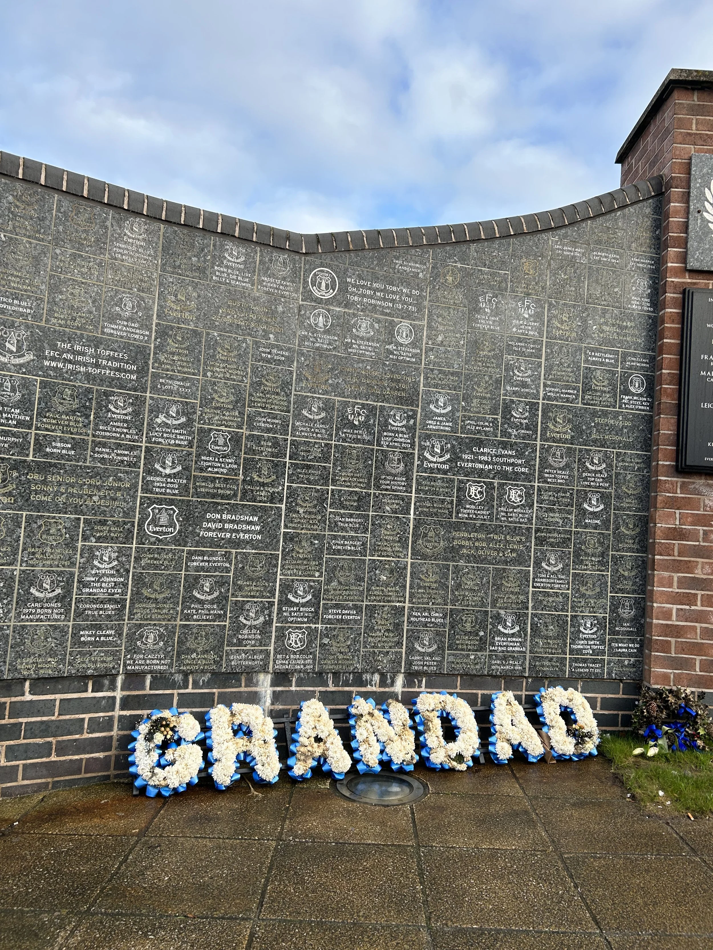 Memorial wall with engraved names, and large floral letters spelling 'GRANDAD' at the bottom, placed on a wet pavement with some flowers nearby.