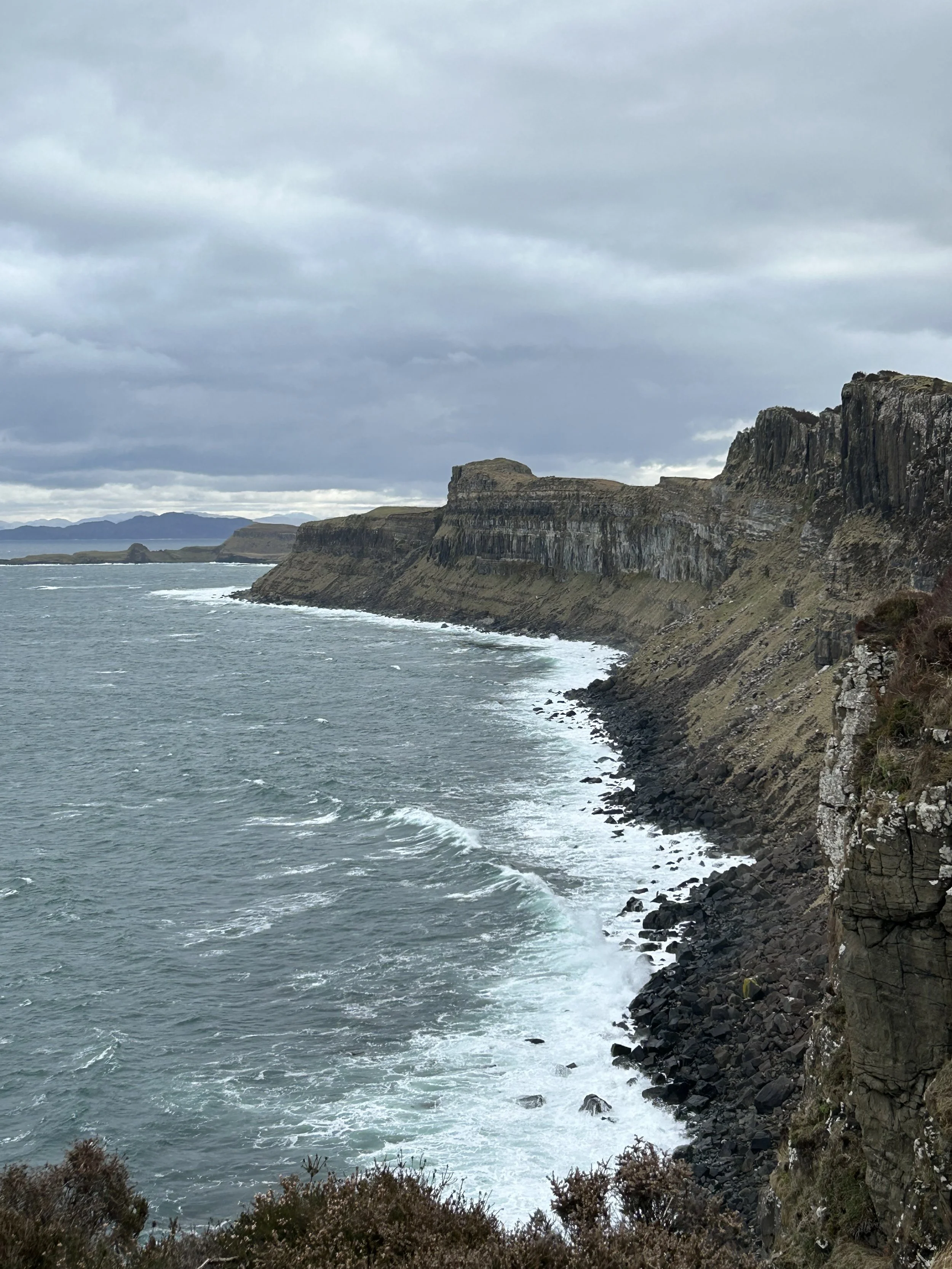 View of rugged cliffs along a coastline with waves crashing against rocks under a cloudy sky.