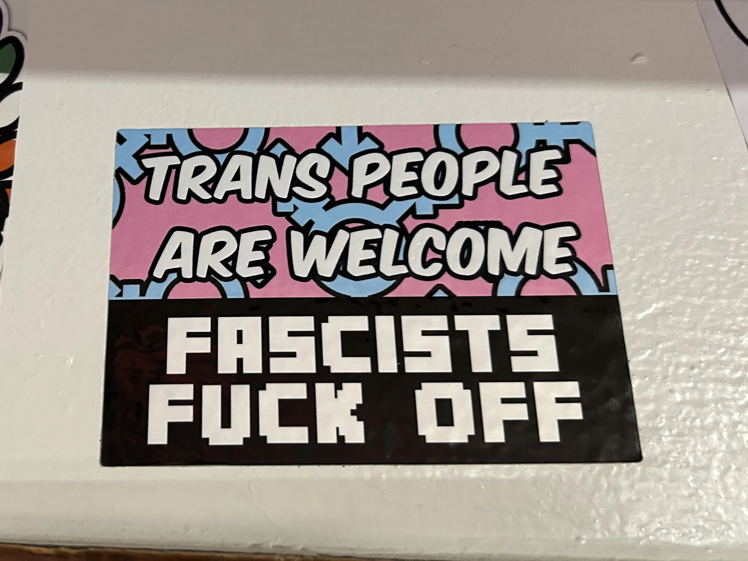 A sticker that says 'Trans people are welcome' in colorful text, with a pixelated font below that reads 'Fascists F*** Off' against a black background.