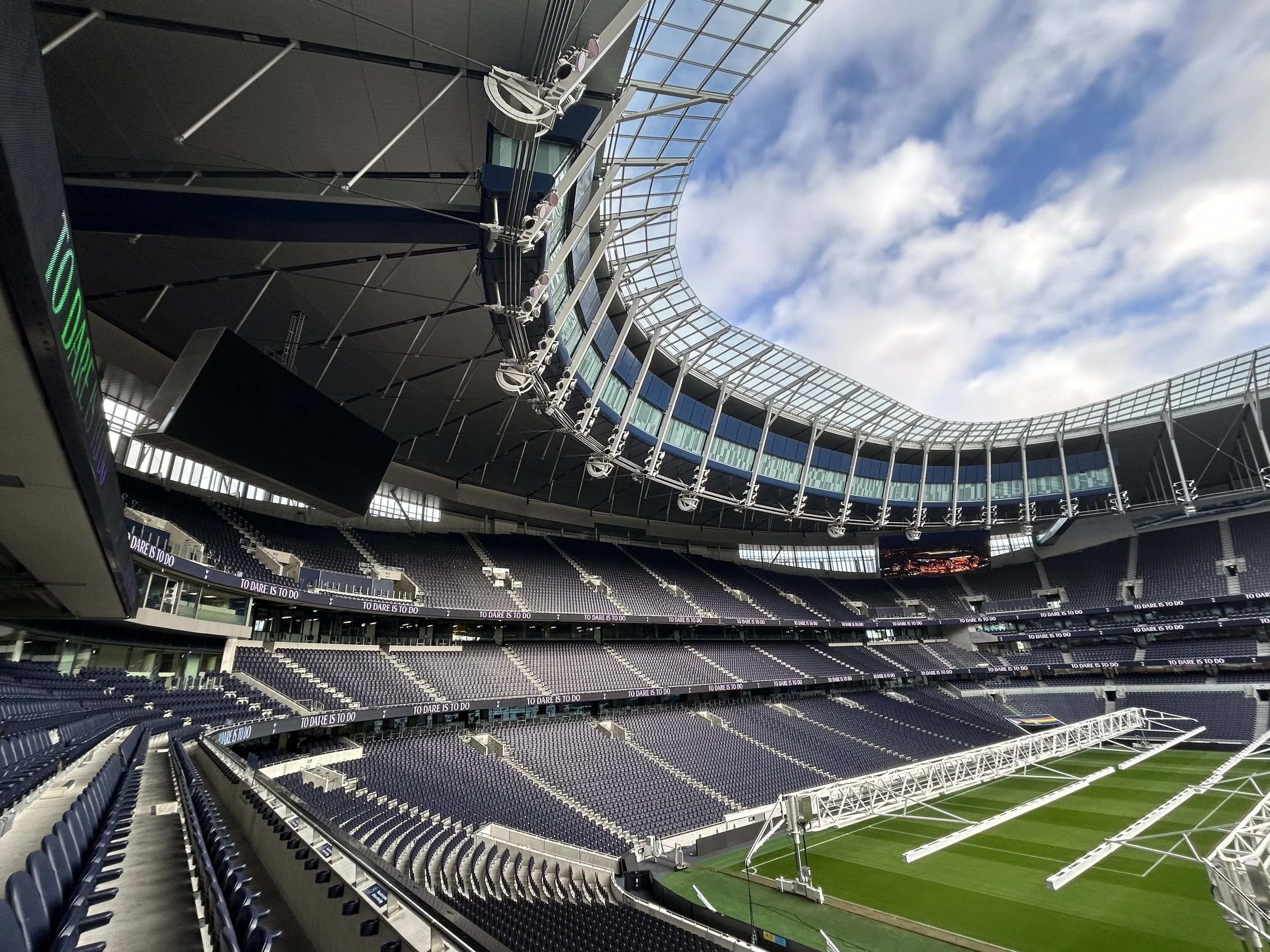 Empty modern stadium with tiered seating and a green field, under a partly cloudy sky.