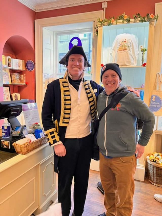 Two men posing inside a shop or cafe, one dressed as a historical military figure with a tall black hat and uniform, the other wearing a beanie and a gray hoodie.