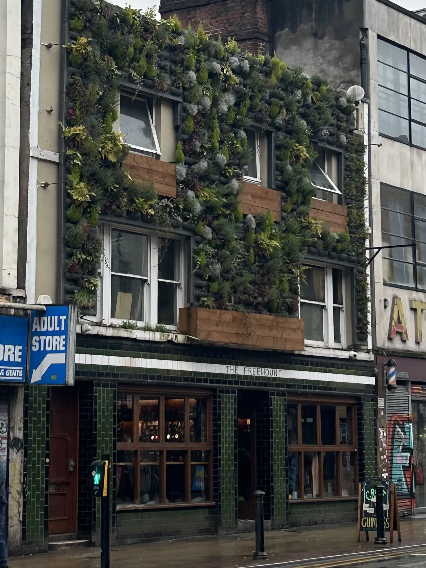 The exterior of a building with a green tile facade and large windows, featuring a vertical garden on the upper levels with various plants and wooden planters. There is a sign for 'The Freemount' and a nearby adult store sign, with graffiti on the ad