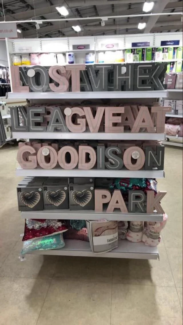 Decorative store display with large letter signs spelling out 'LOVE ATTACK,' 'LEAVE A,' 'GIVE AT,' 'GOOD IS ON,' and 'PARK,' with Christmas decorations and household items on shelves.