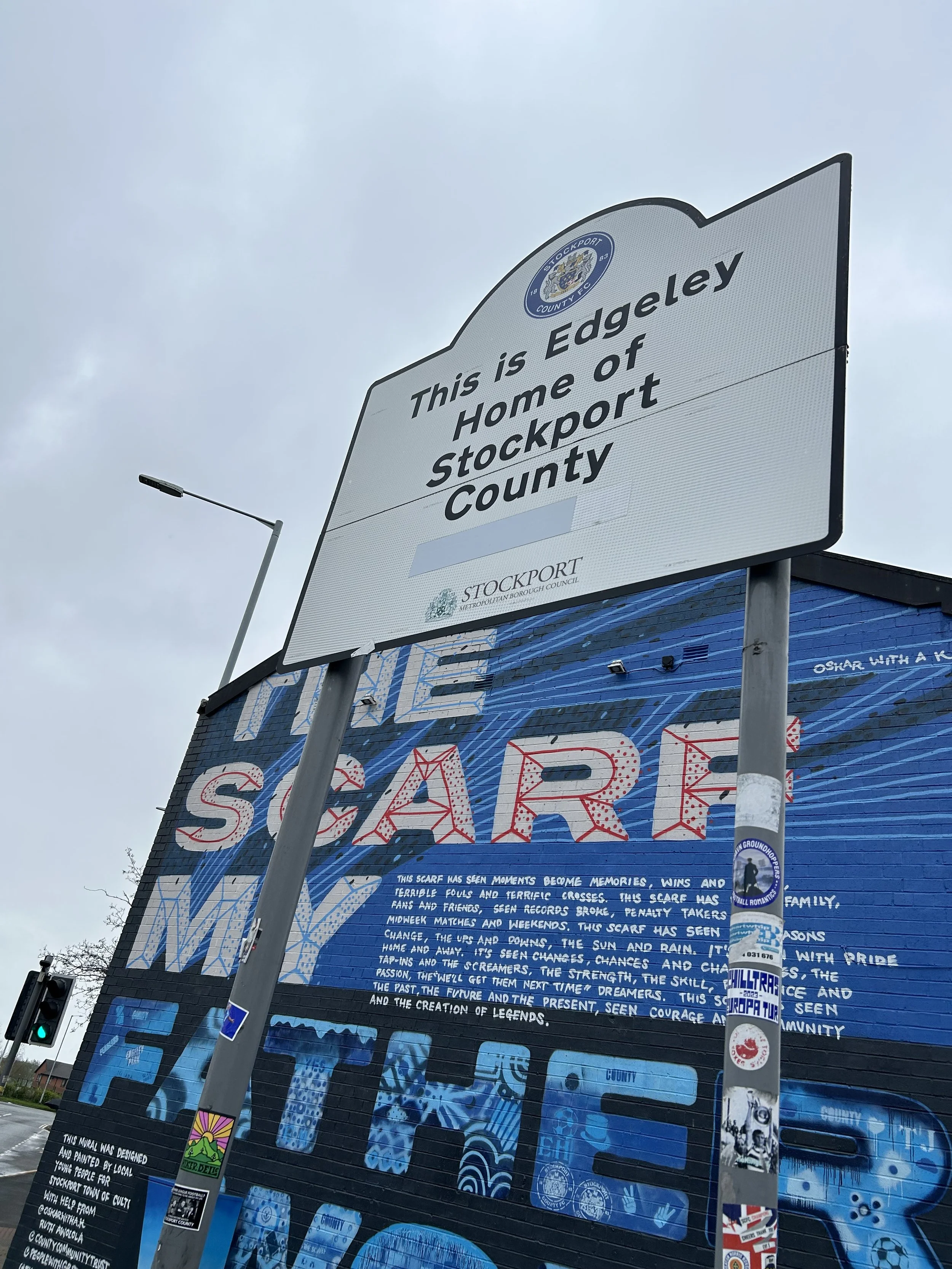 A street view with a large sign that says, 'This is Edgeley, Home of Stockport County,' with a smaller sign below indicating it is in Stockport Metropolitan Borough Council. Behind the sign, there is a painted mural on a building with colorful letter