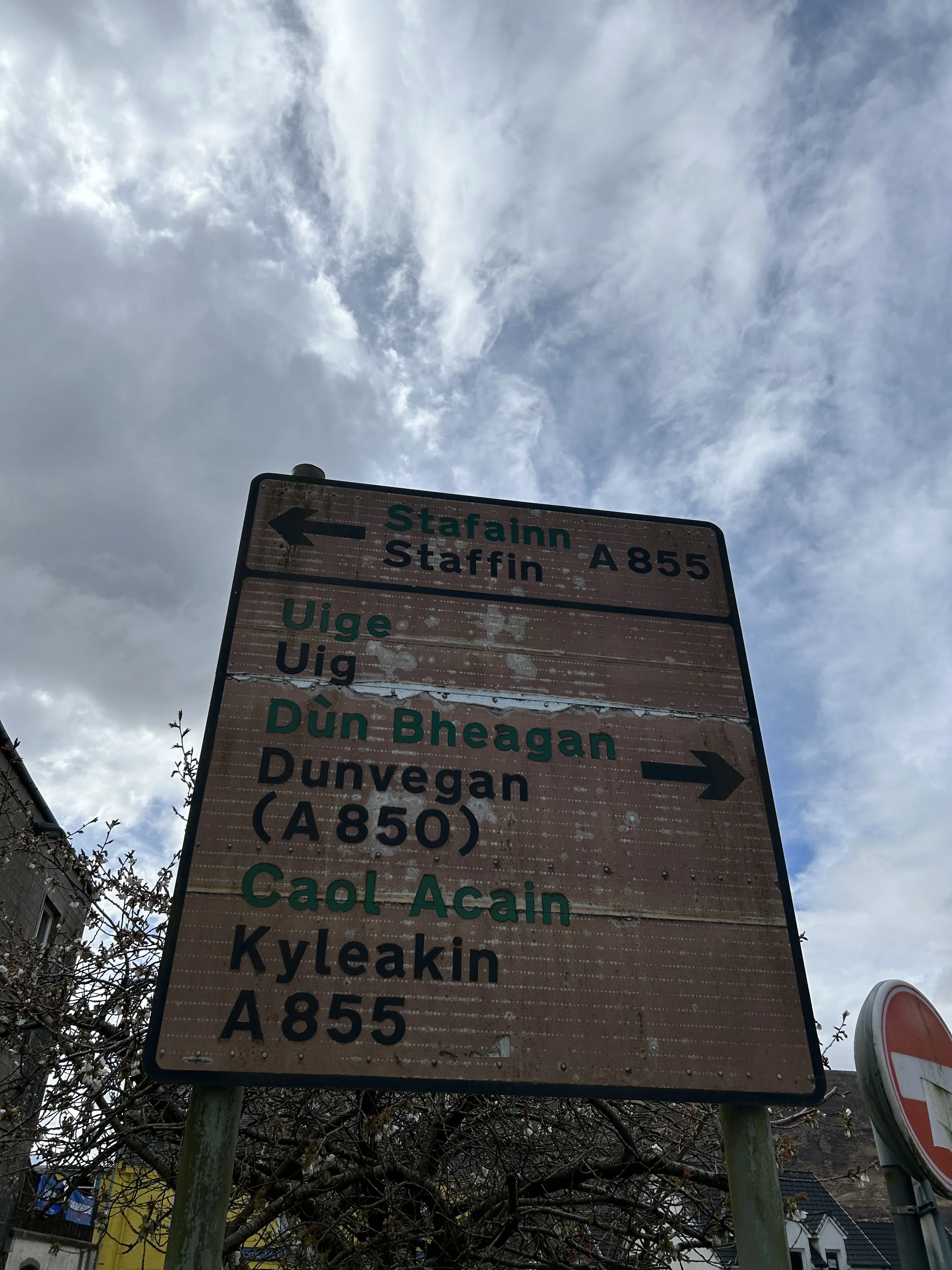 Weathered road sign showing directions to Staffinn A855 to the left and Dunvegan A850 to the right, with other locations listed below. Overcast sky in the background.