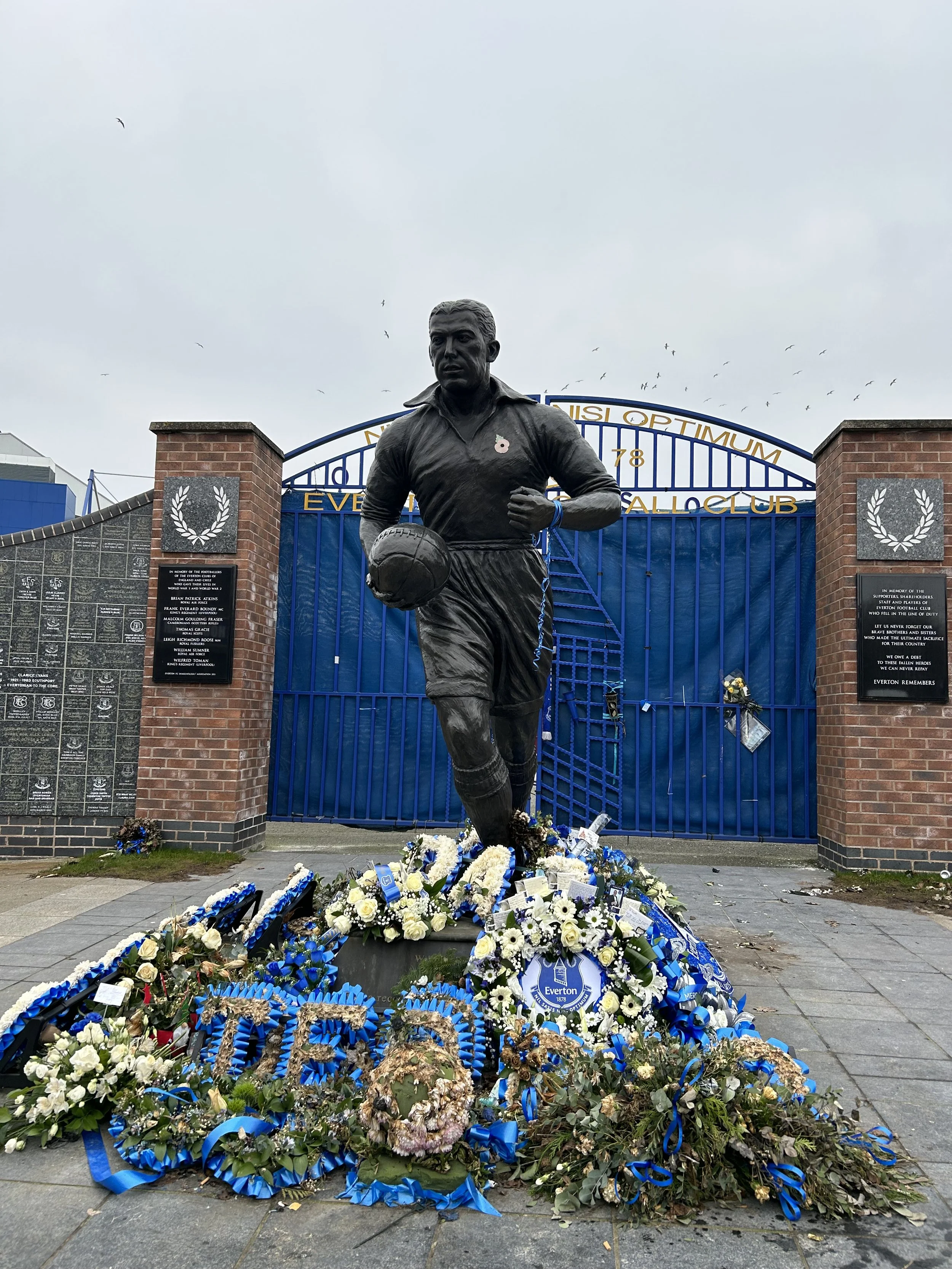 Statue of a football player holding a ball in front of a blue gate with floral wreaths and commemorative plaques at Everton Football Club in memory of victims of the 1989 Hillsborough disaster.
