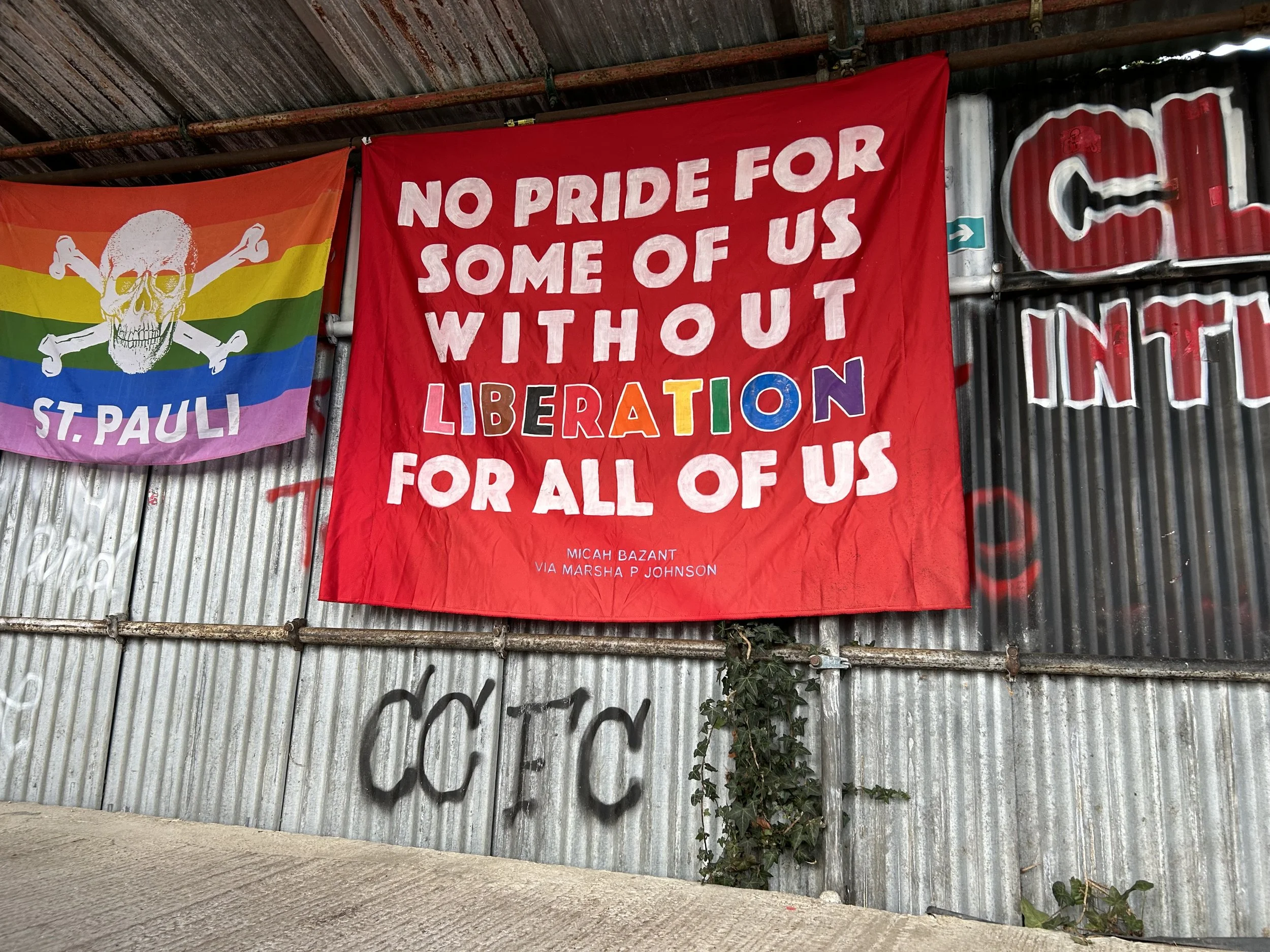 Red banner with white and multicolored text that reads, "No pride for some of us without liberation for all of us." The name Micah Bazant and Marsha P. Johnson are written at the bottom. To the left, a rainbow flag with a skull and crossbones in the 