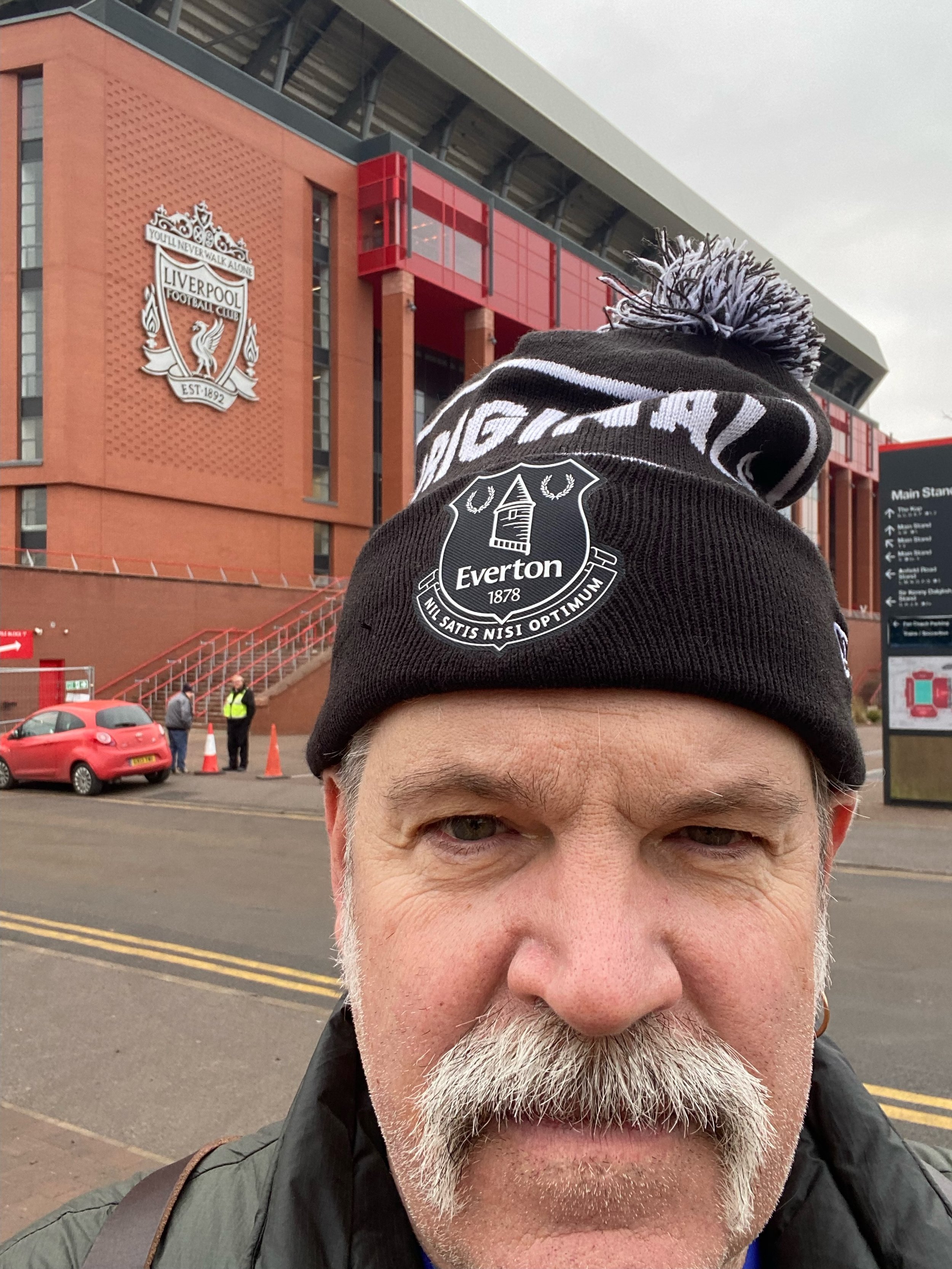 A man wearing a black Everton beanie hat taking a selfie outside of Liverpool FC stadium. The stadium and signs are visible in the background.