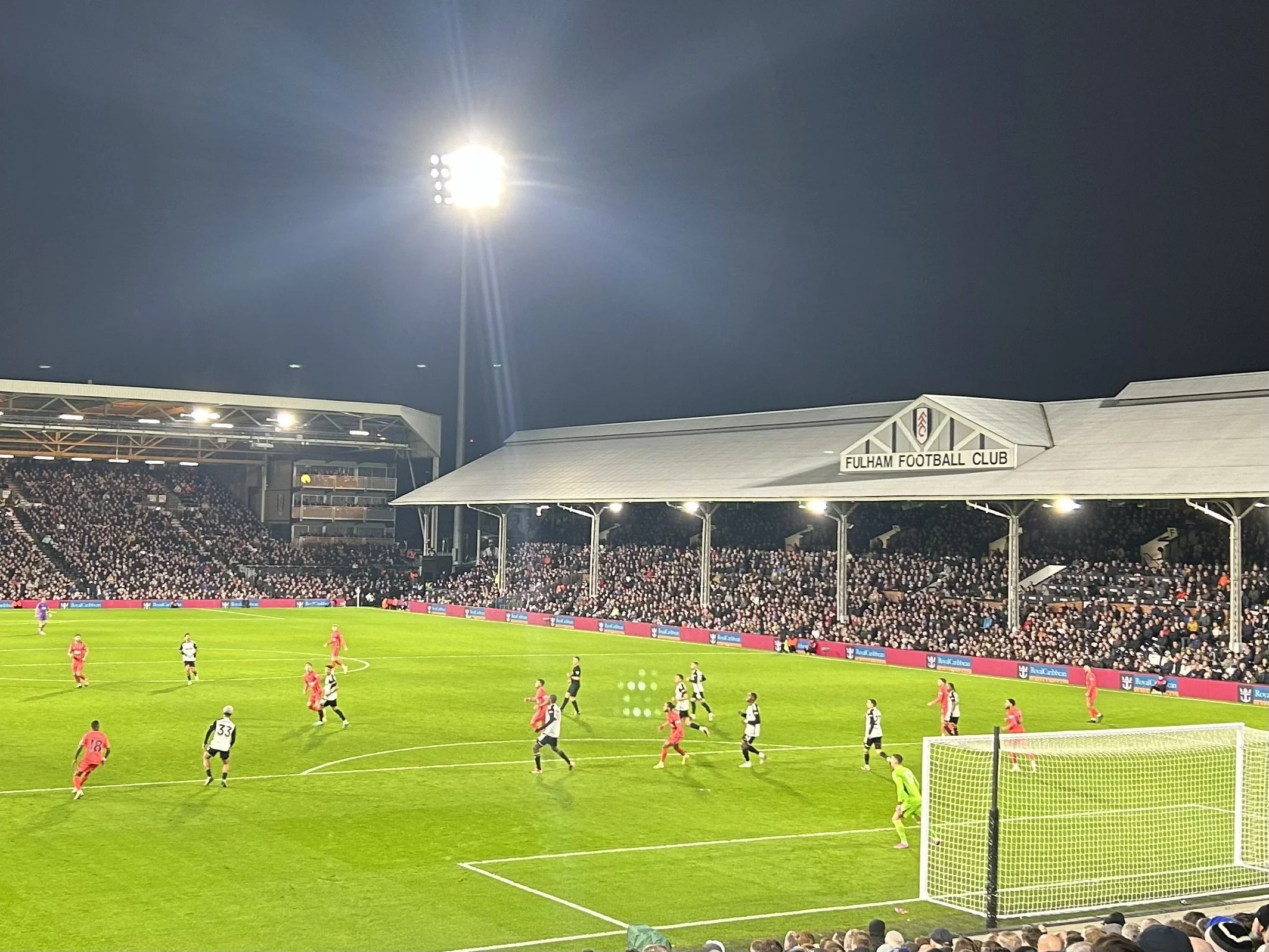 A soccer match at Fulham Football Club stadium at night, with players on the field, spectators in the stands, and bright stadium lights illuminating the scene.