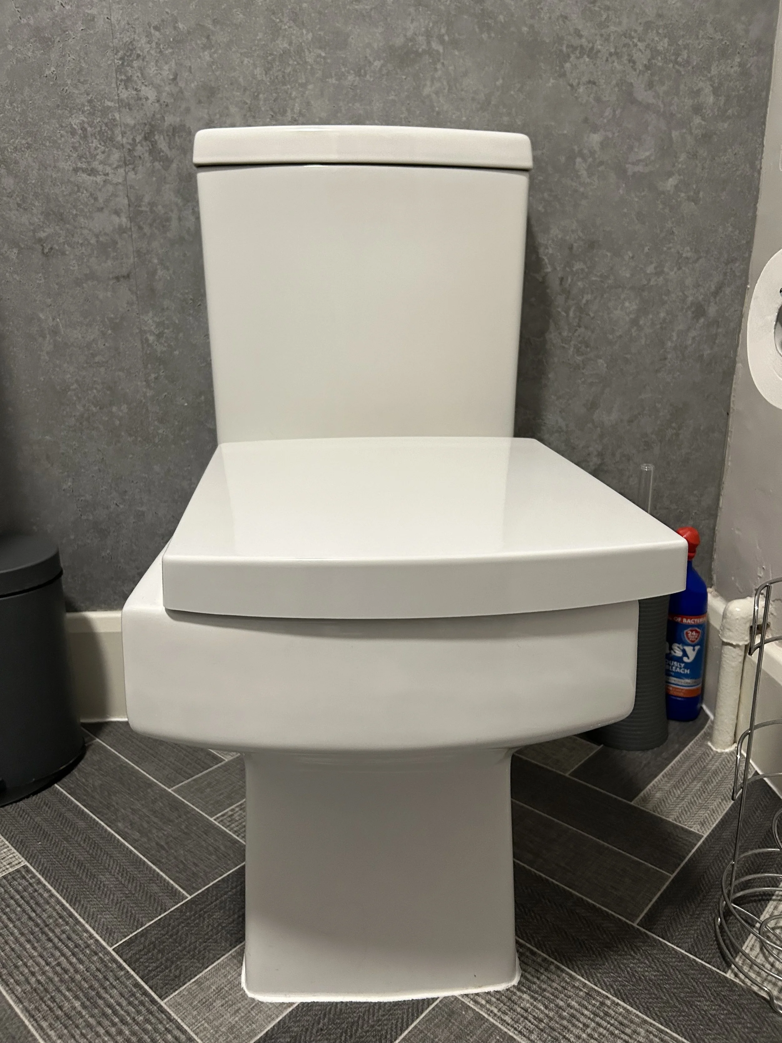 A modern white toilet in a bathroom with gray tiled walls and dark gray floor tiles.