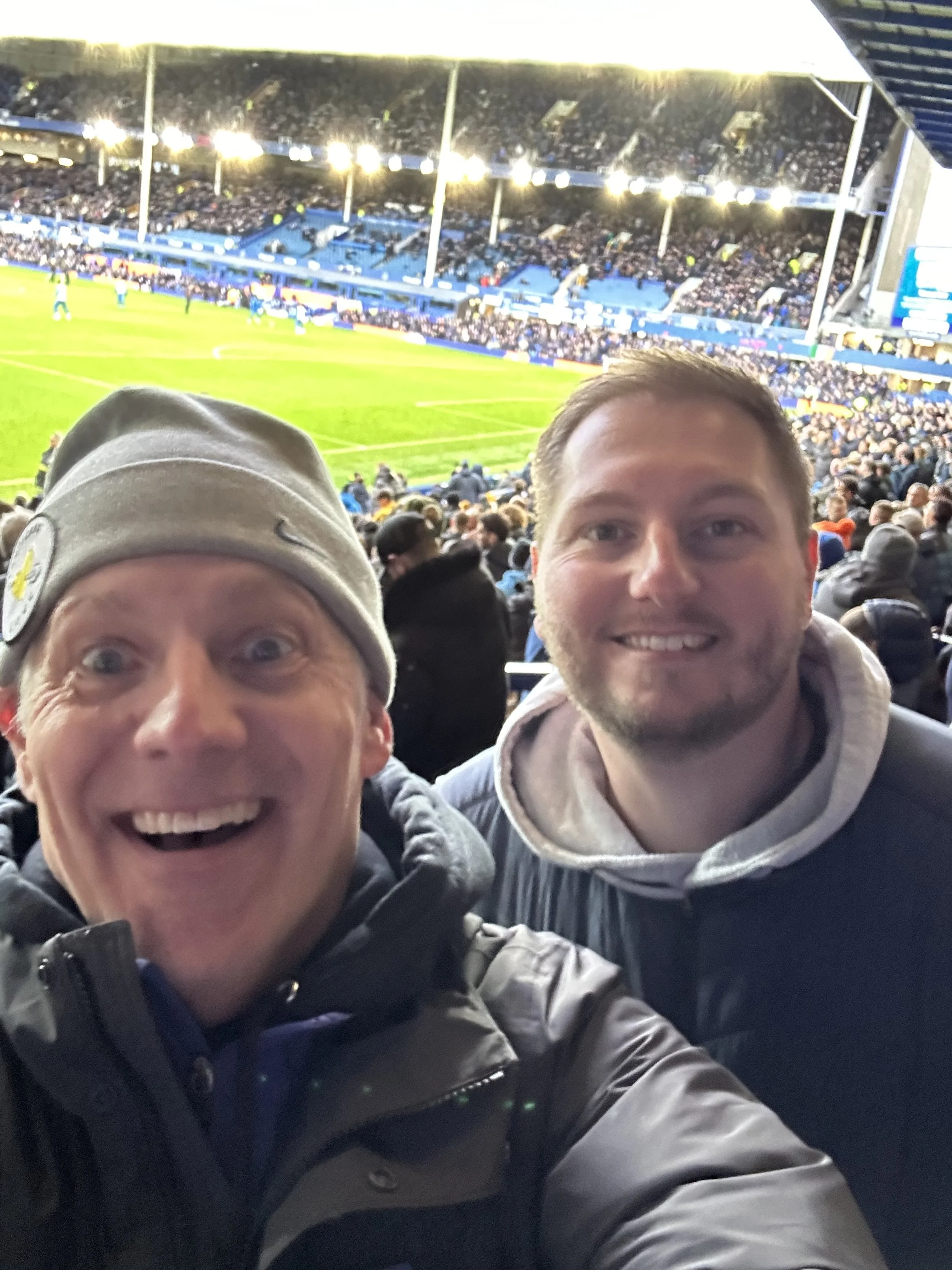 Two men smiling at a sports stadium during a football game, with a crowd and field in the background.