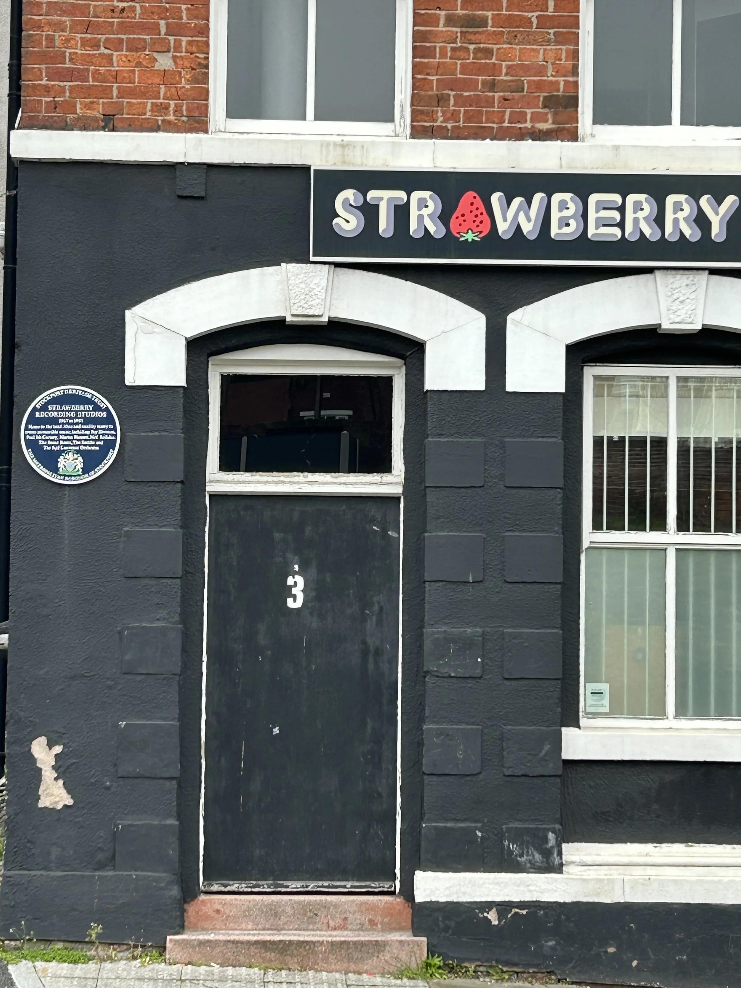 Black building with white accents, a window, door marked number 3, and a sign that reads 'Strawberry' with a strawberry illustration.
