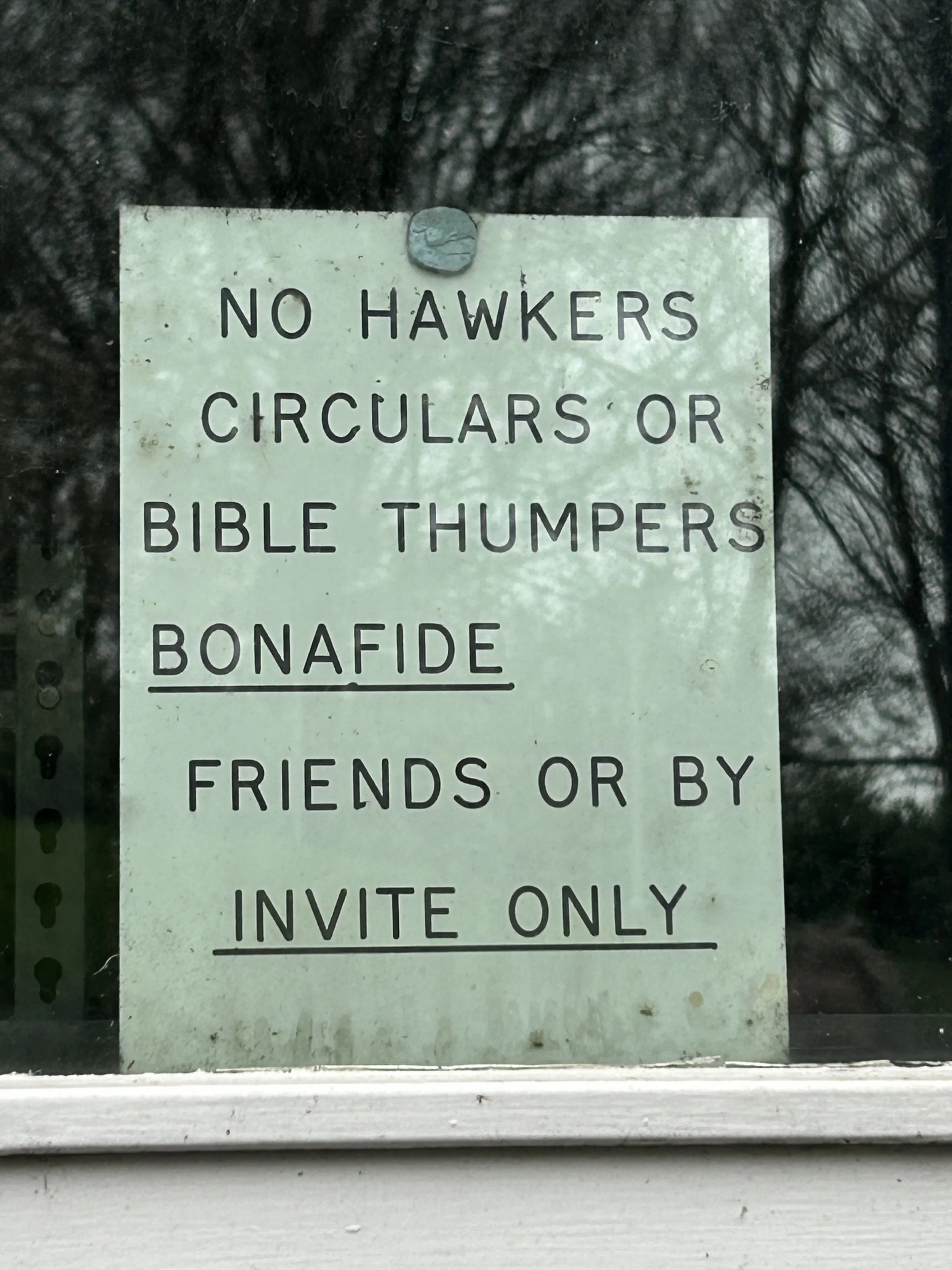 Sign with rules against hawkers, circle or Bible thumpers, bonafide friends or by invite only, posted on a window with trees reflected in the background.