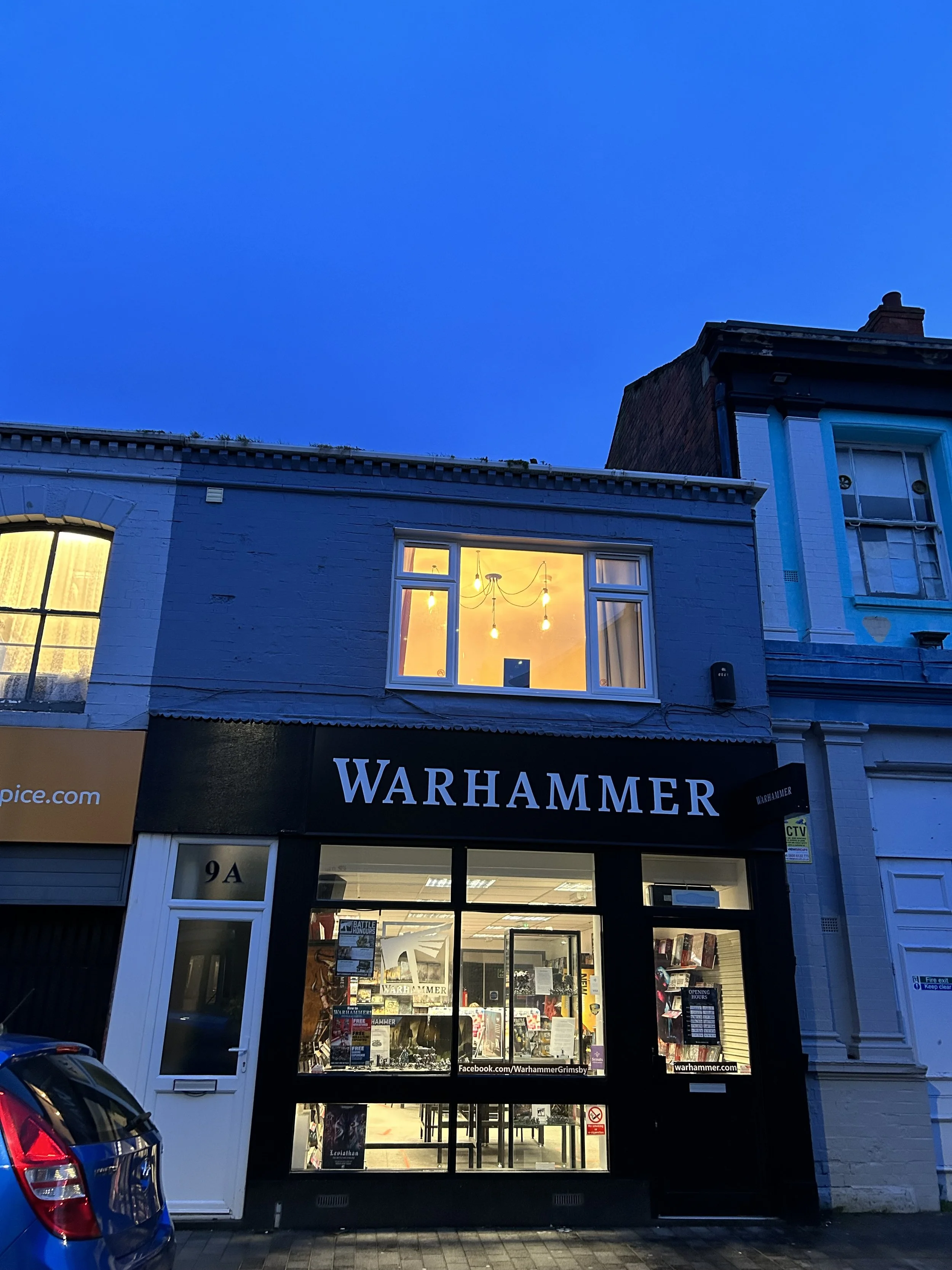 Street view of a building with a gaming store called Warhammer, illuminated interior visible through large front window, blue sky in the background.