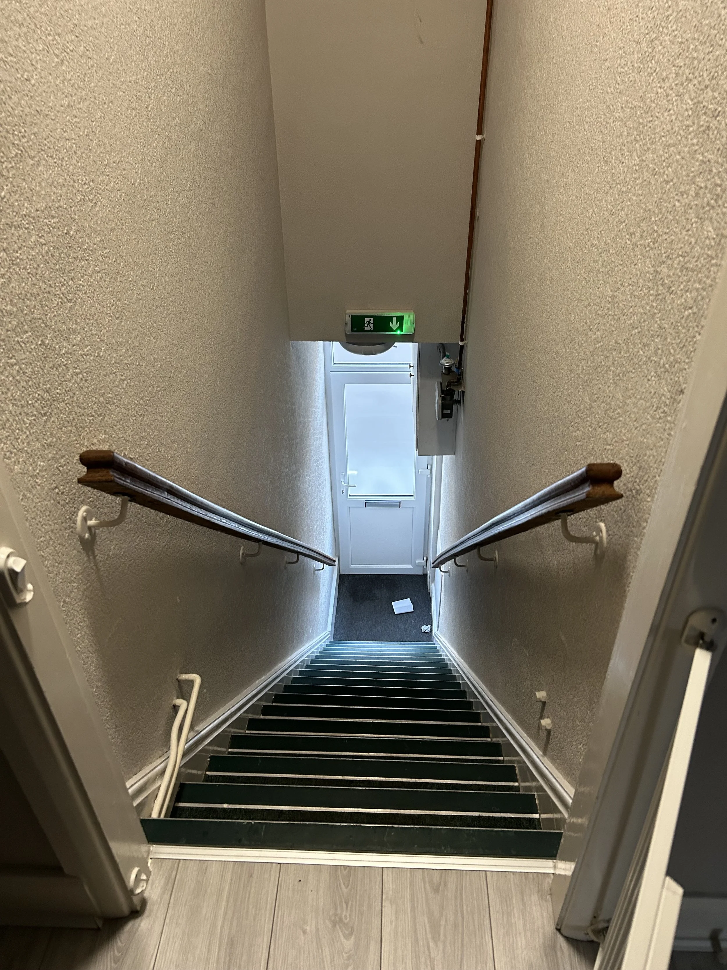 Looking down a staircase from the top, with handrails on both sides, leading to a door with a glass window and an exit sign above it. There is a small white box and some trash on the floor at the bottom of the stairs, and a water fountain is visible 