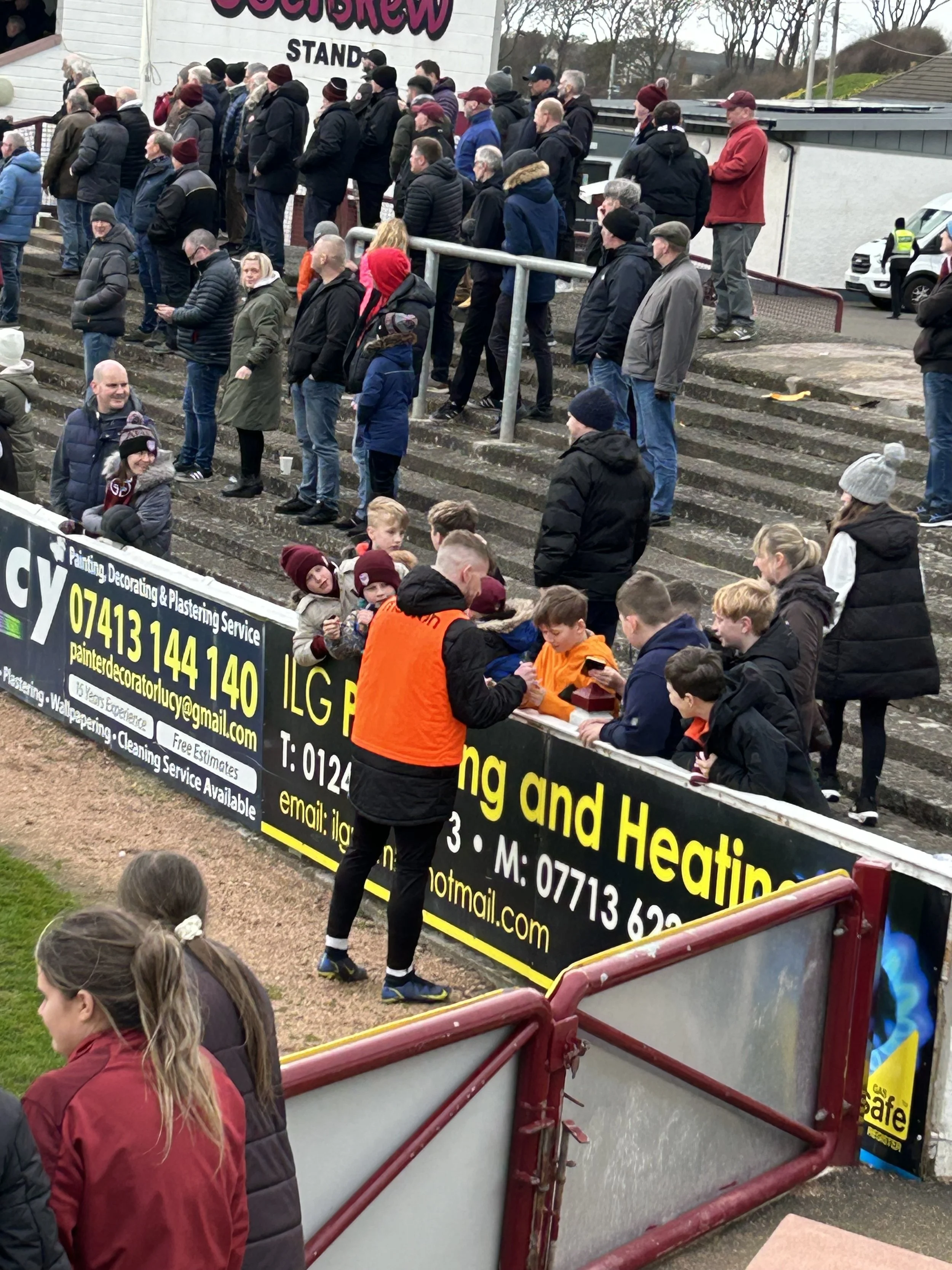 A crowd of people waiting in line at a football stadium entrance on a cloudy day, with some individuals wearing jackets and hats, and a man in an orange vest interacting with children at a ticket or registration table near advertising boards.