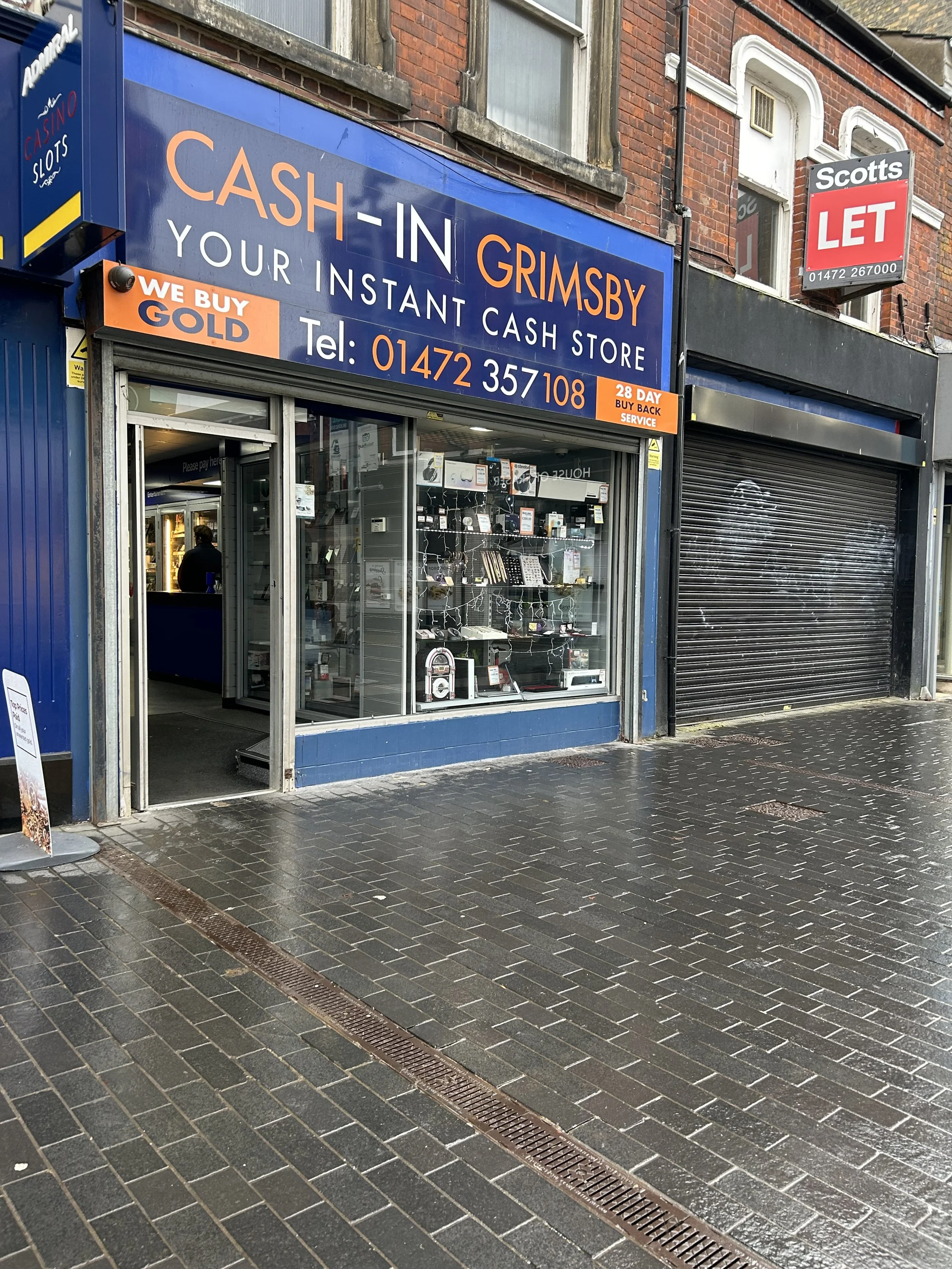  storefront signage for 'Cash-In Grimsby,' a cash store with a blue and orange sign, phone number 01472 357108, and a slogan 'We buy gold.' The store has a glass window display with various electronics and accessories inside, located next to a black 