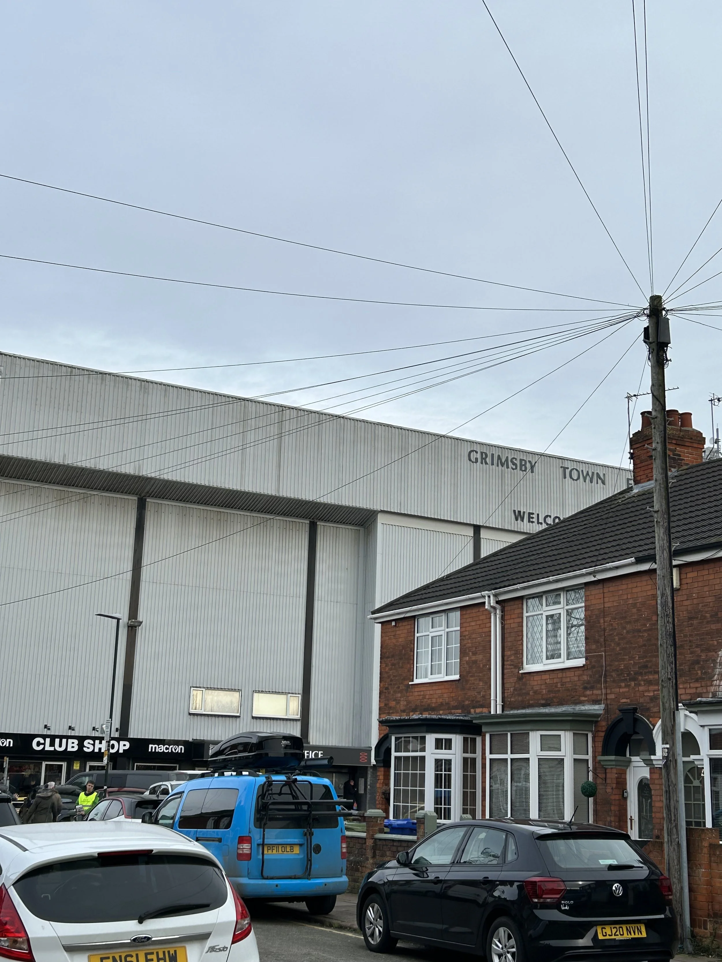 Street view of a residential block with a brick house, parked cars, a utility pole, and a large building in the background with the sign 'Grimsby Town'.