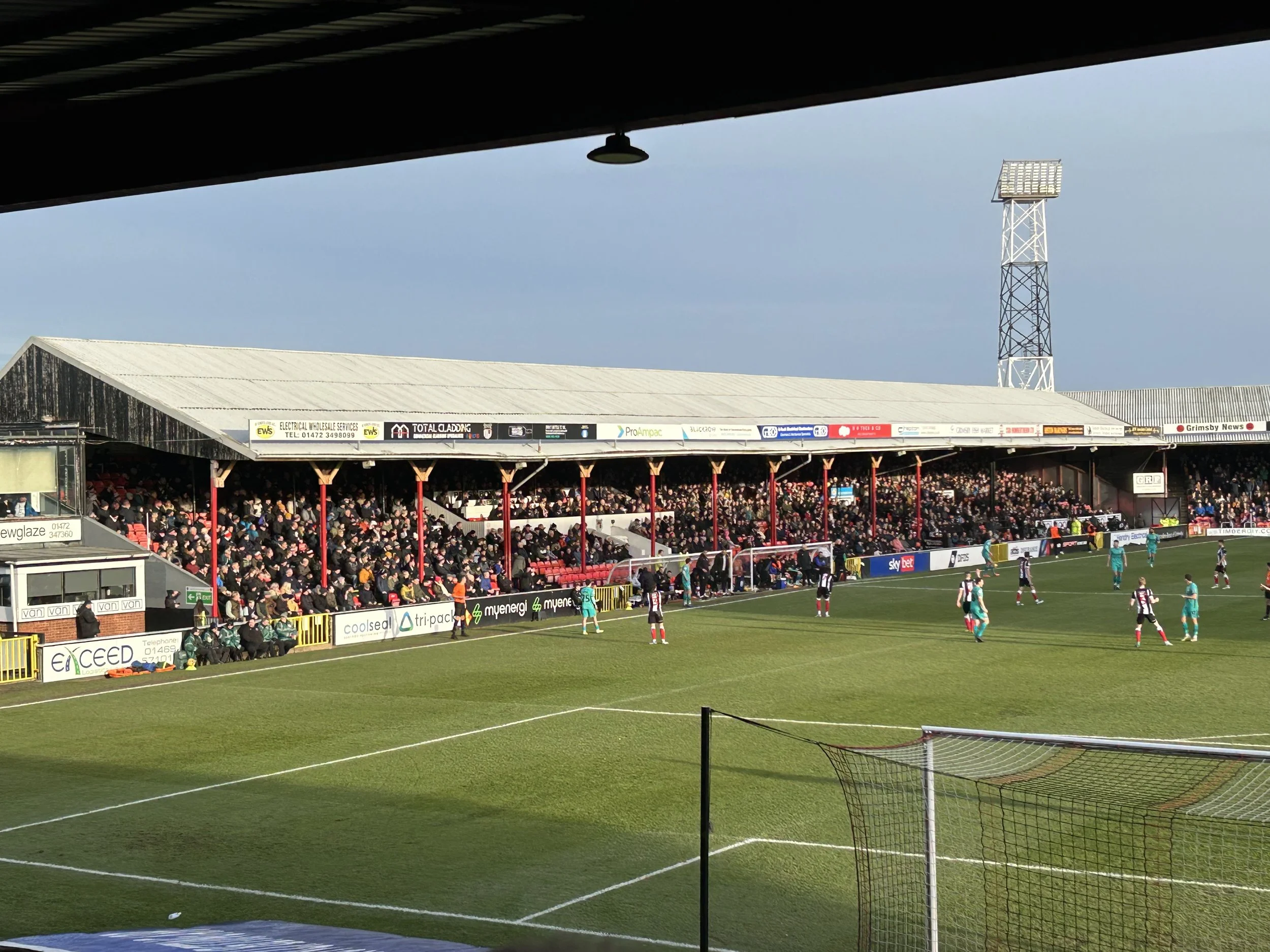 A football match in a stadium with players on the field, spectators in the stands, and a large floodlight tower in the background.