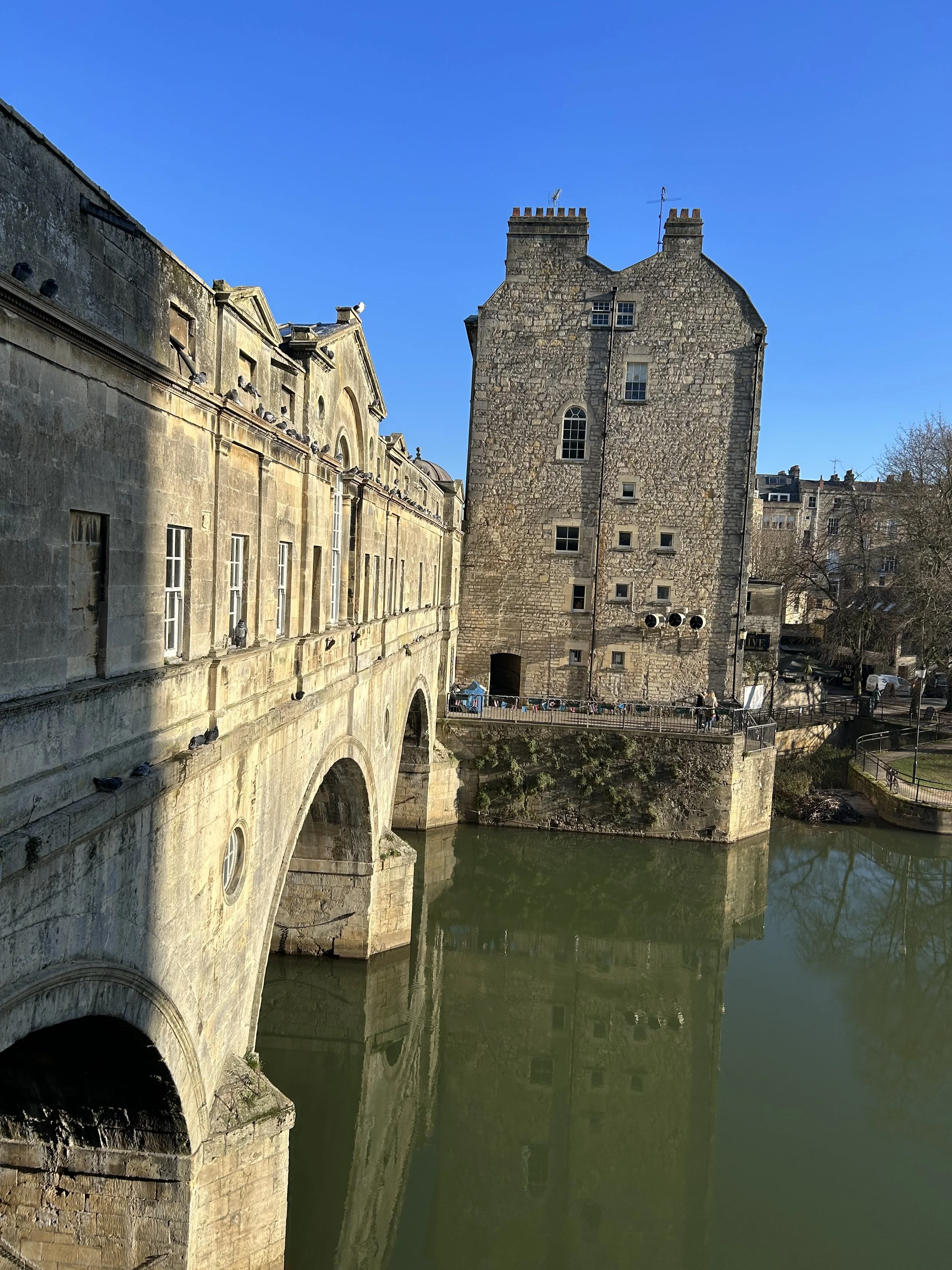 Historic stone building with arched windows and a tower next to a river, under a clear blue sky.