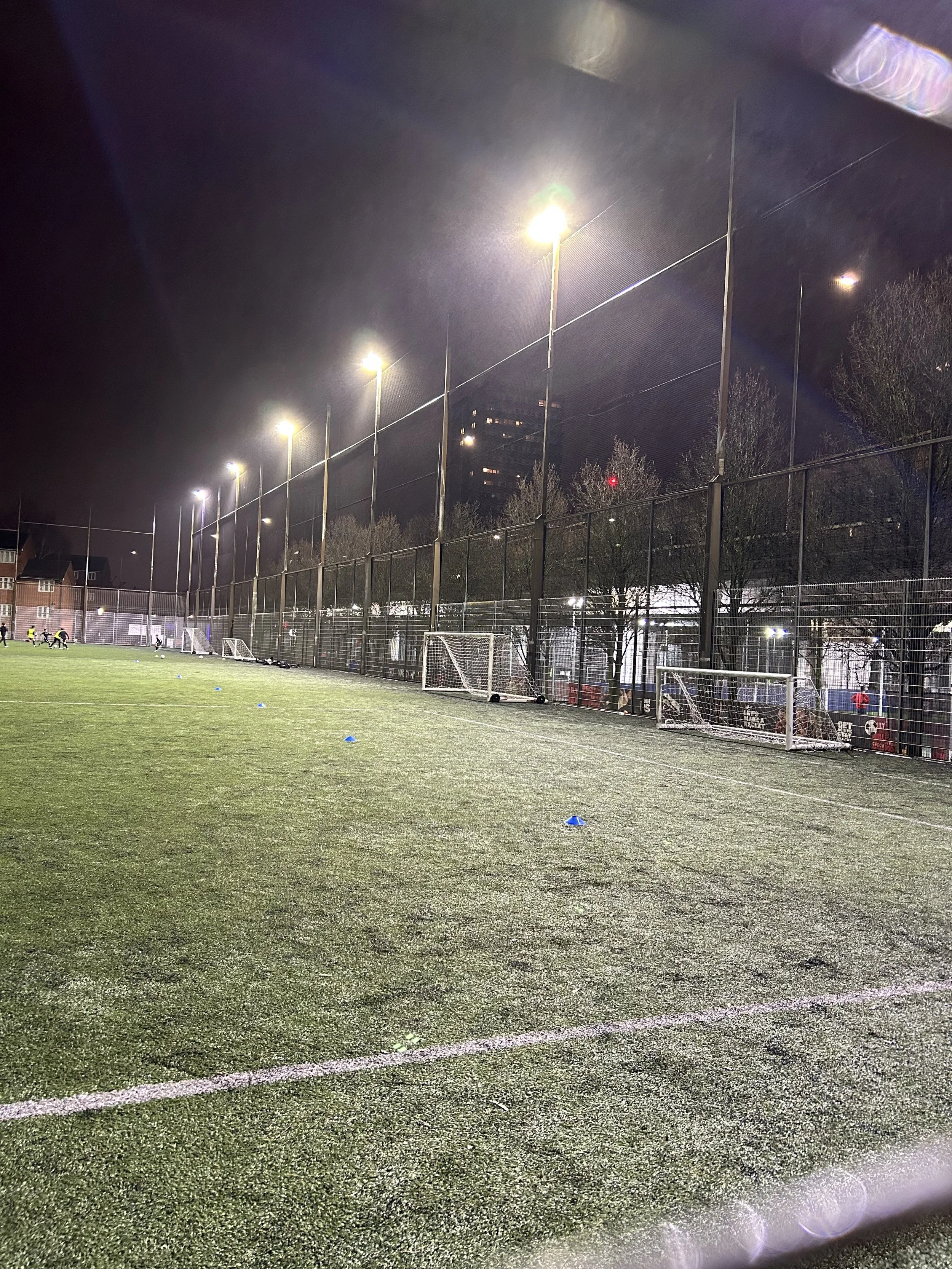 Nighttime view of an outdoor soccer field illuminated by tall lights, with a chain-link fence, small soccer goals, and children playing in the distance.