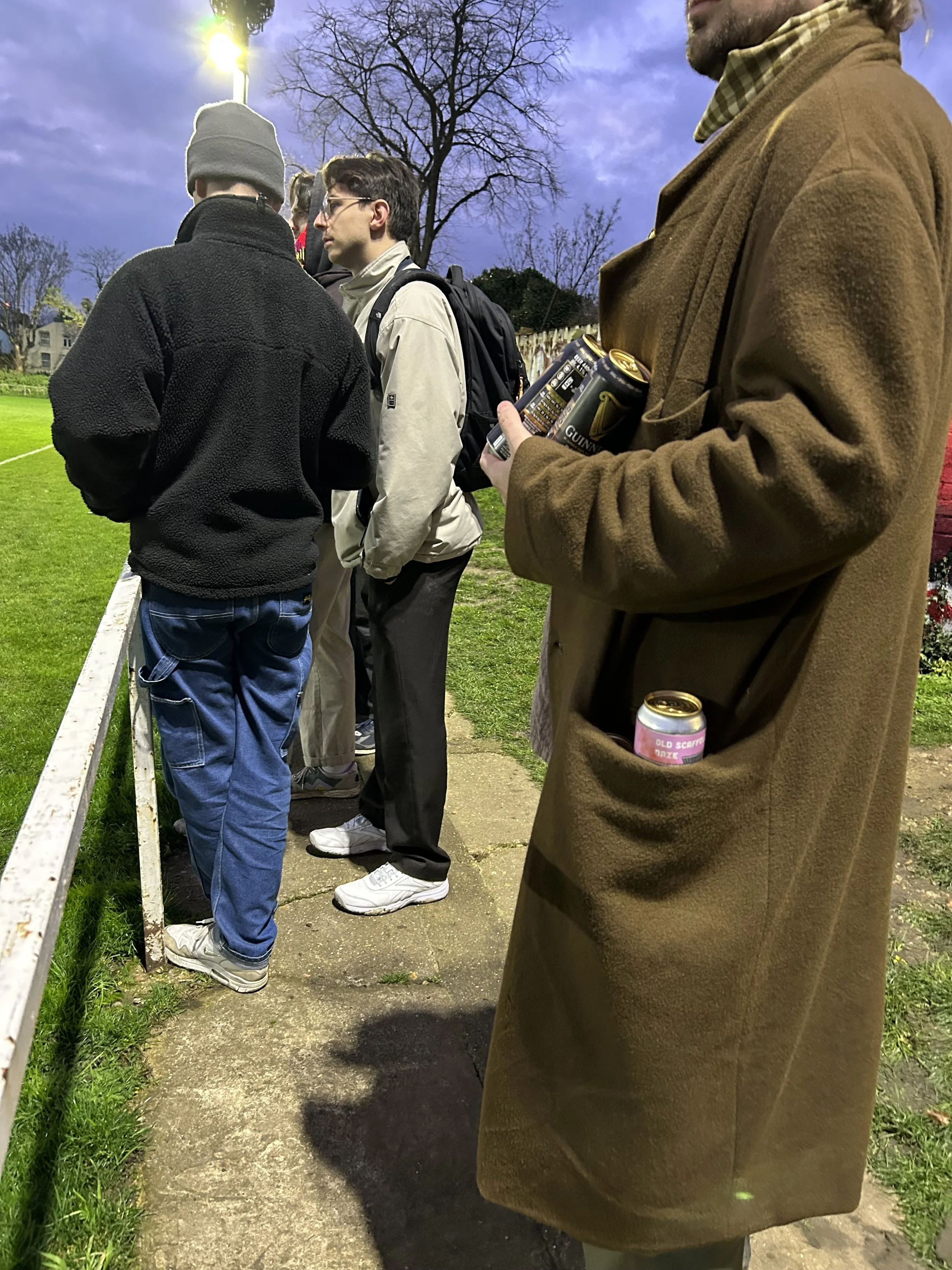 Group of people standing outdoors on a paved path, some holding cans of Guinness beer, with grassy field and trees in the background during dusk.