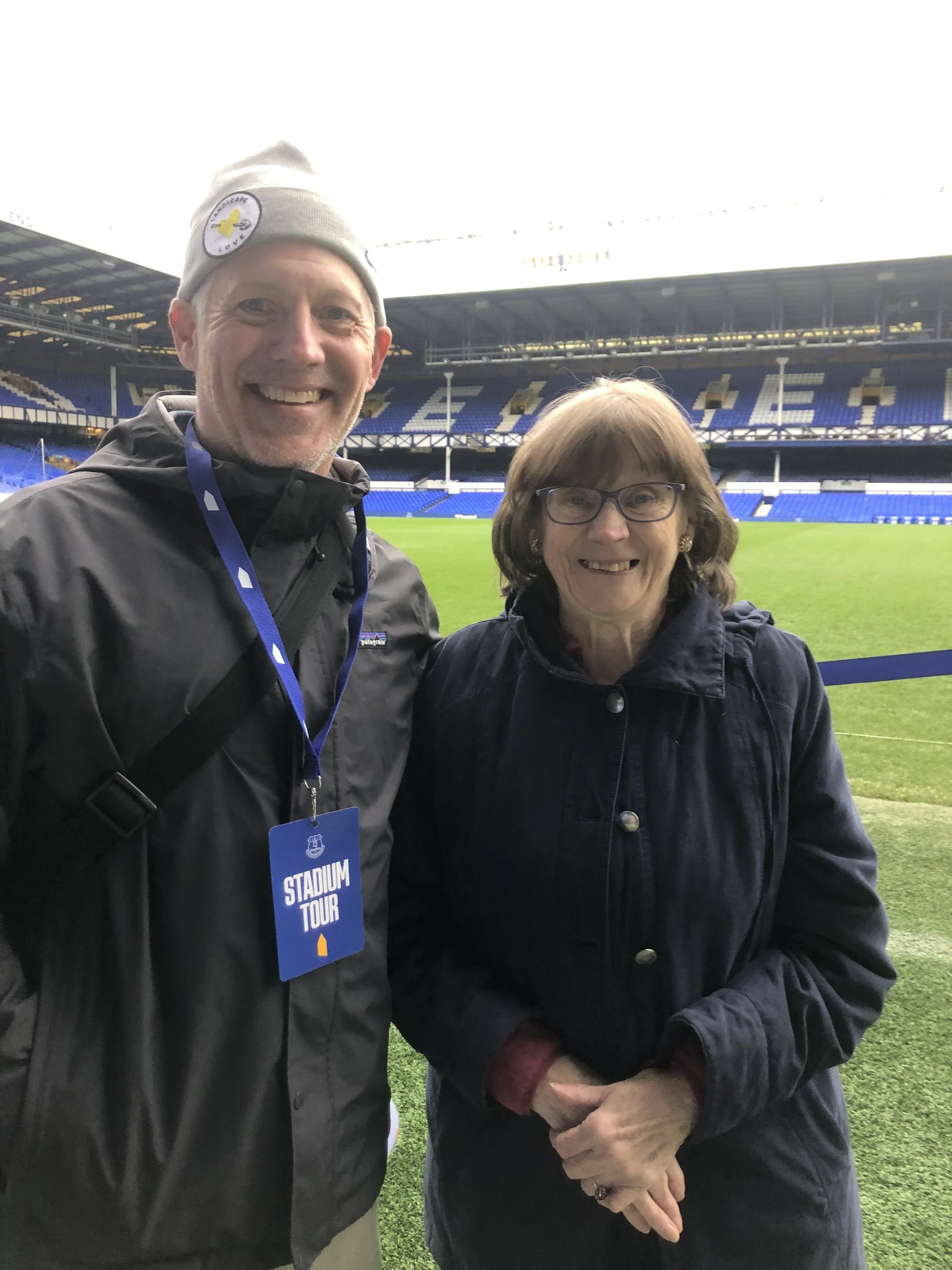 A man and woman smiling at a football stadium with empty blue seats and green field in the background.